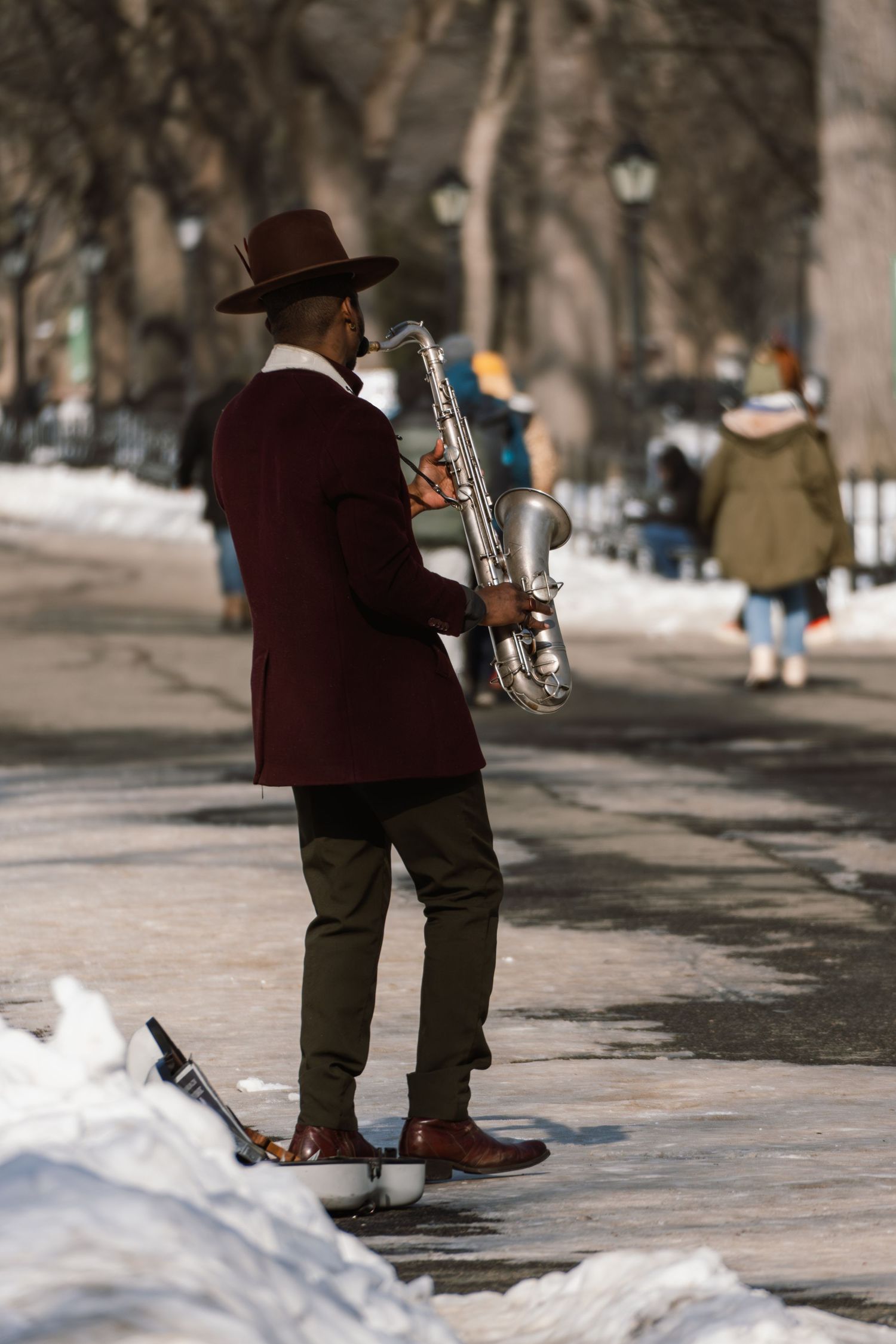 A saxophone player in a brown hat and burgundy jacket performs outdoors on a snowy winter day.