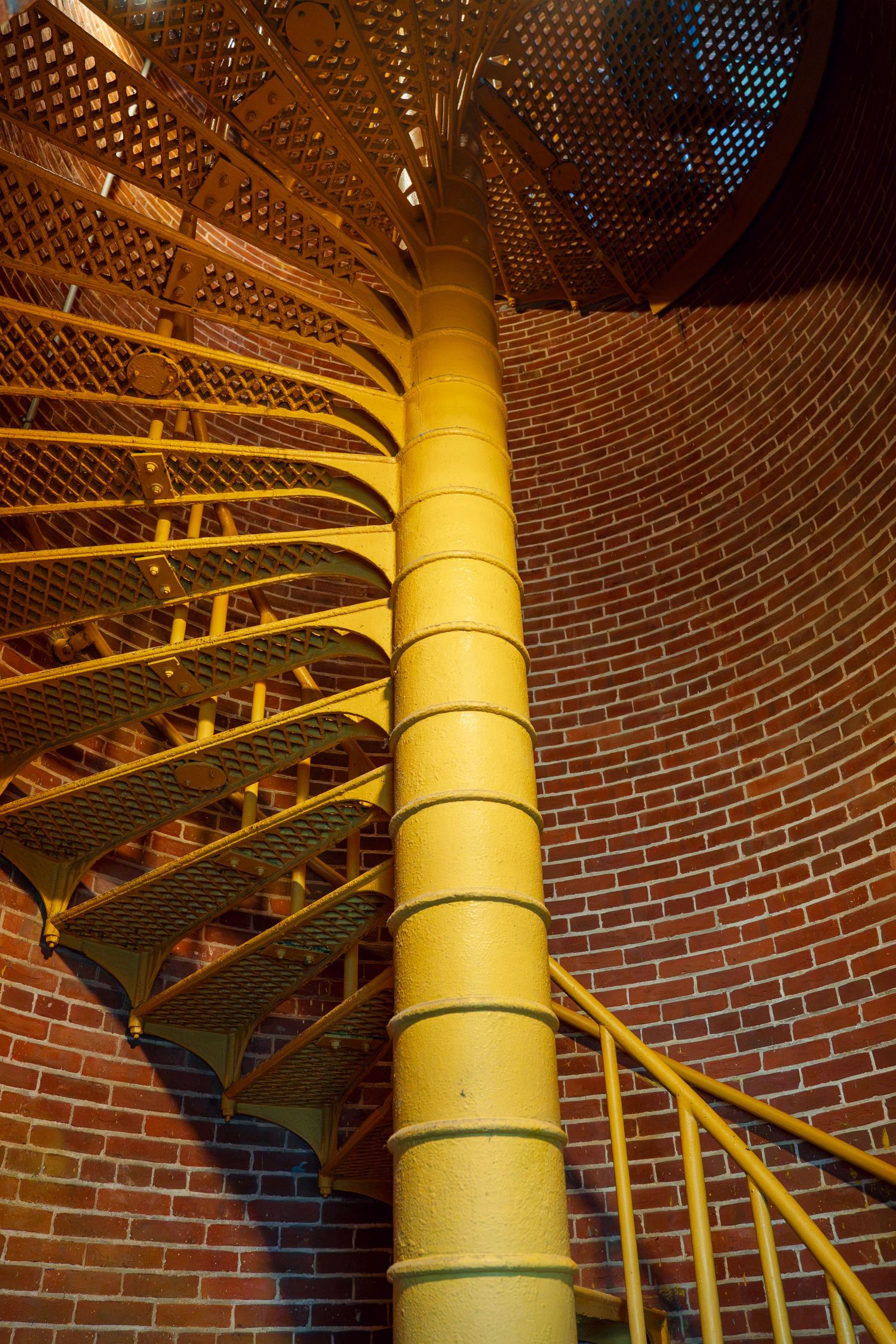 Yellow spiral staircase with metal steps winds around a central pole inside a brick lighthouse tower.