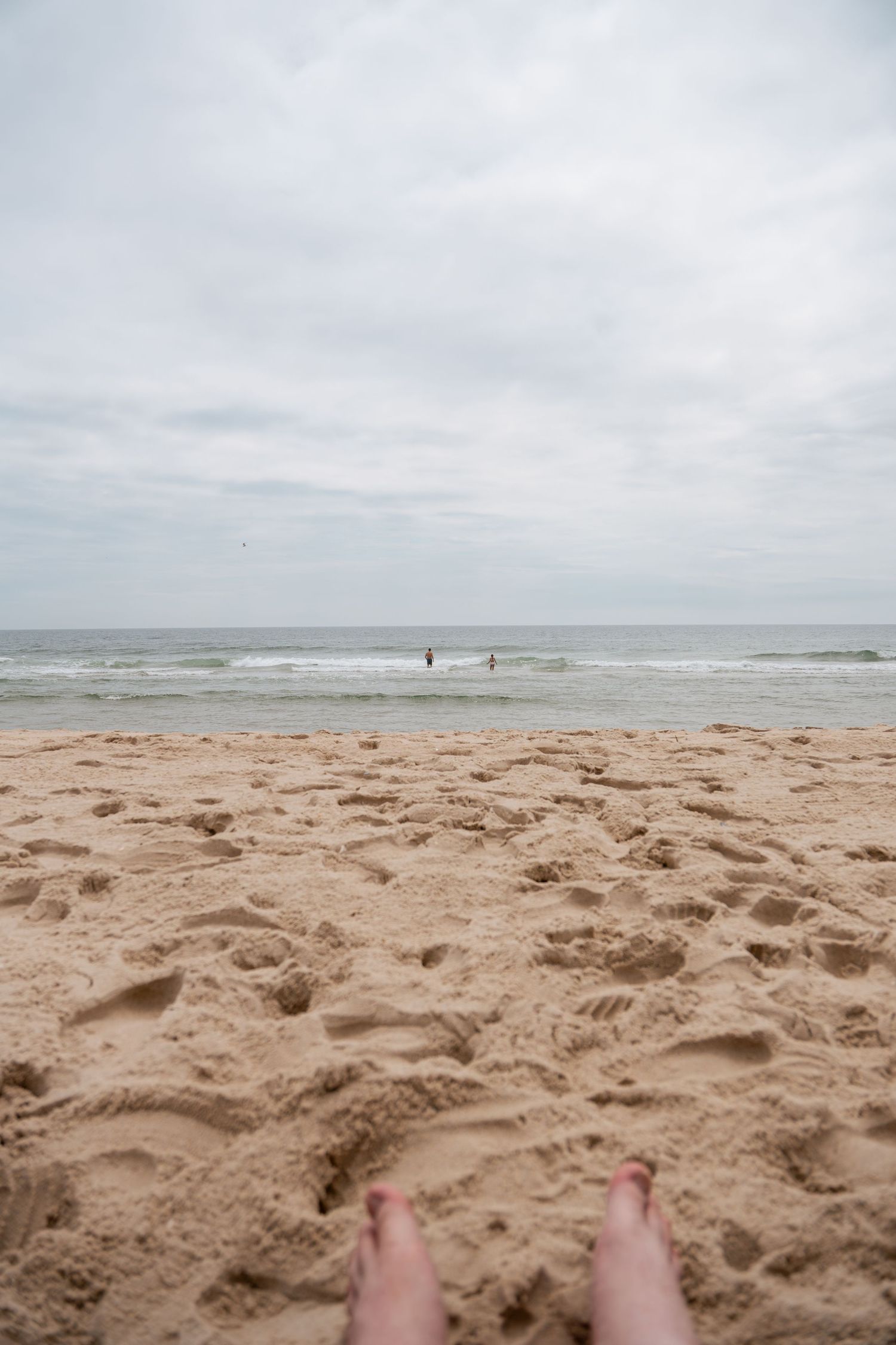 Person relaxing on a sandy beach with footprints, calm ocean waves, and overcast sky in the background.