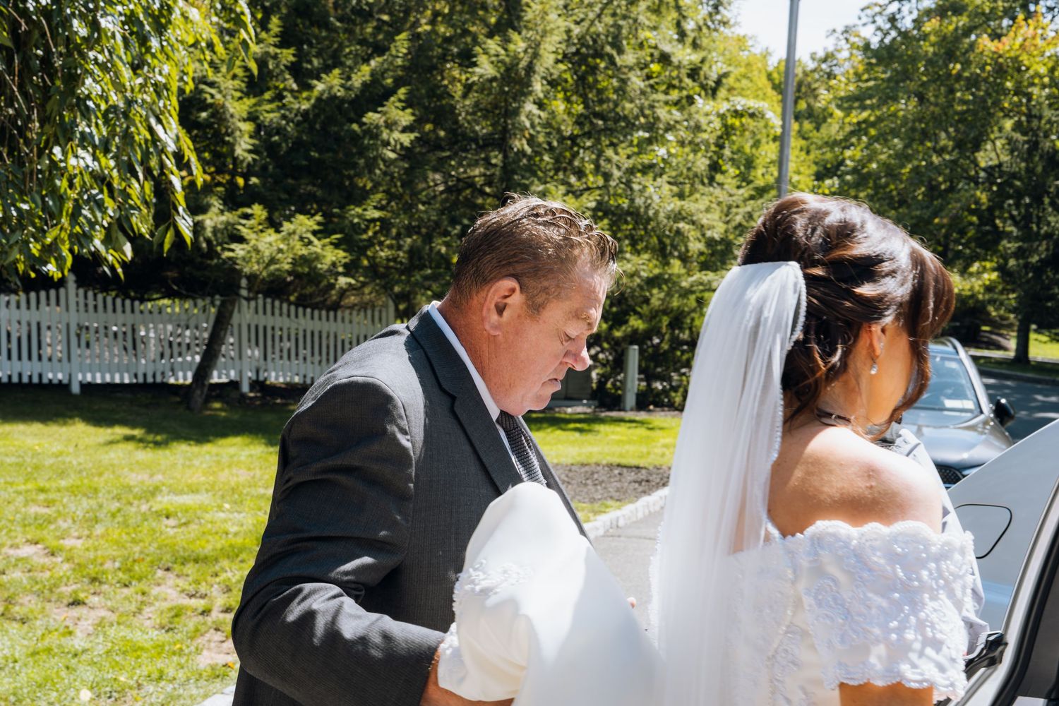 A groom in a gray suit helps a bride in a white lace dress and veil near a car on a sunny wedding day.