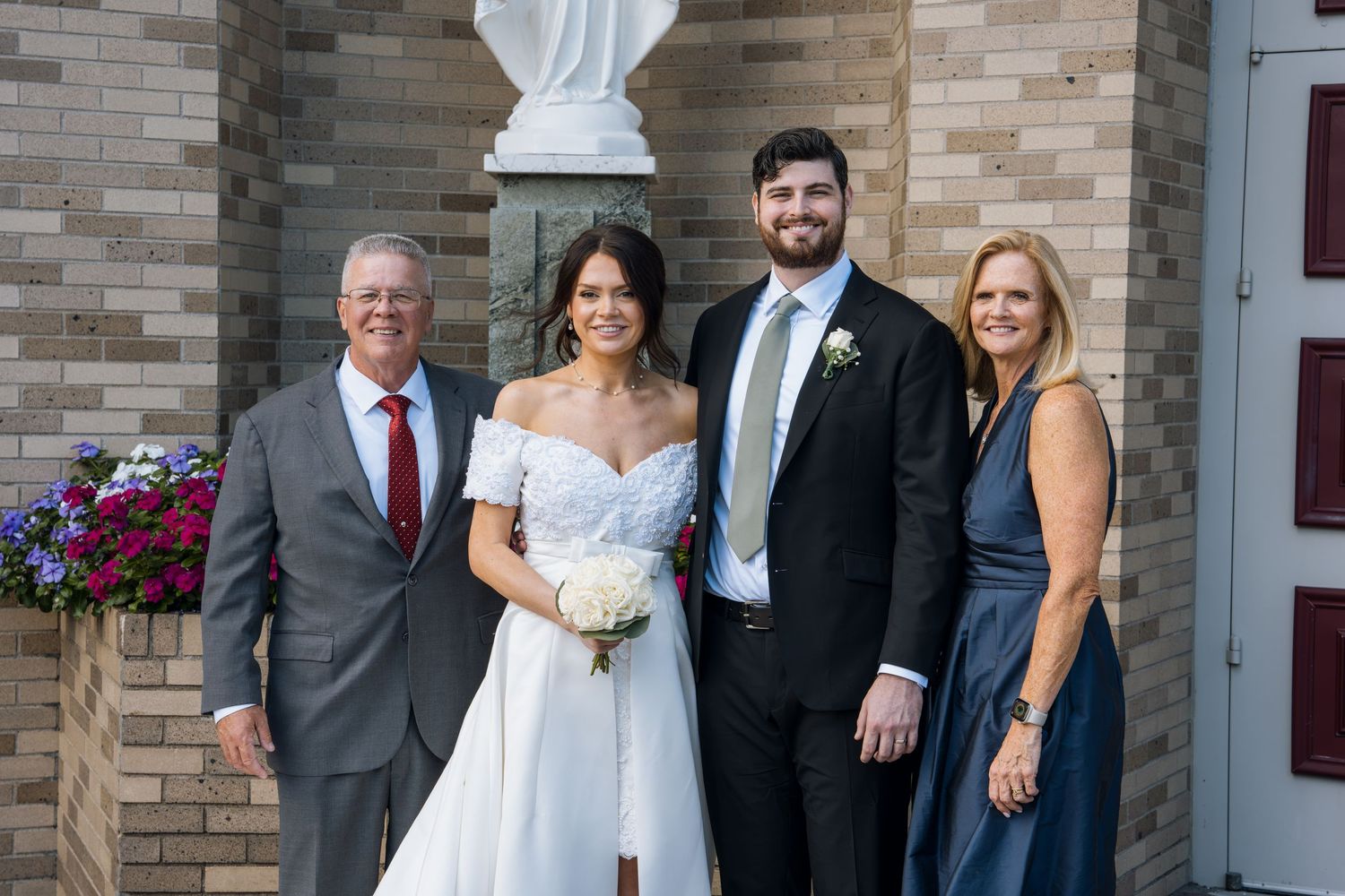 Wedding party of four posing outside a brick church, bride in white gown holding bouquet with groom and family.