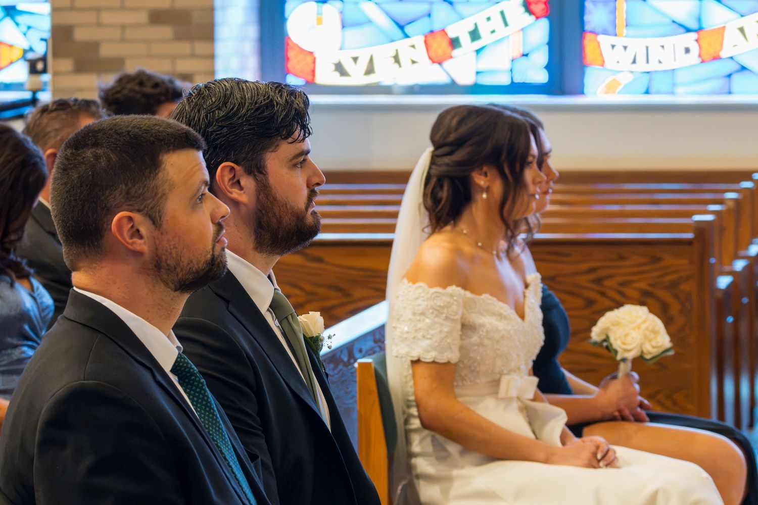 Bride in white off-shoulder gown and groom in suit seated at church wedding ceremony with stained glass windows.