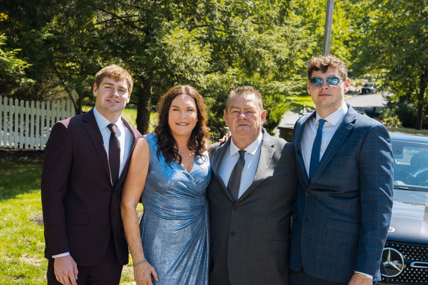 Family of four posing outdoors in formal attire, smiling together at a sunny event near a Mercedes vehicle.