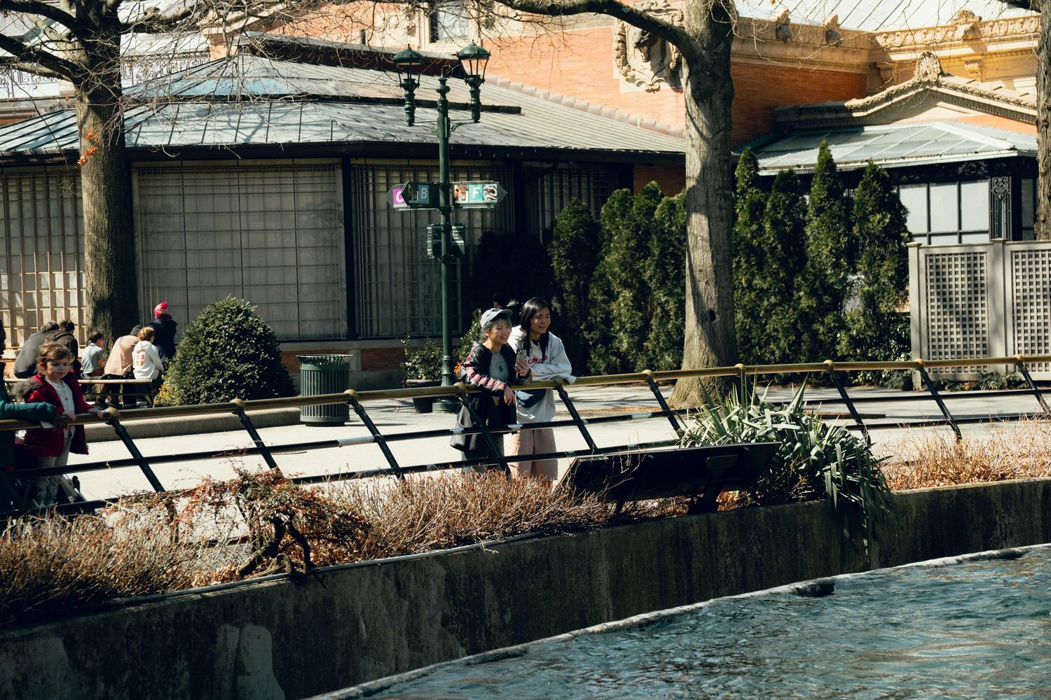 People walking along a scenic canal path lined with bare trees and traditional Japanese buildings.