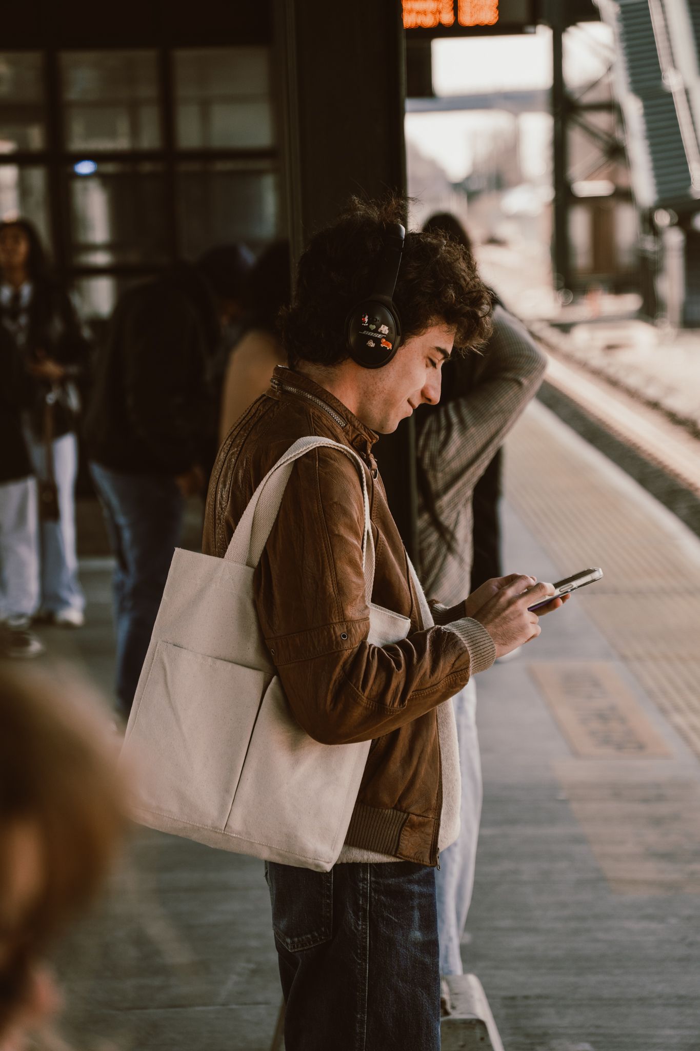 Young man wearing headphones and carrying a tote bag looking at his phone while waiting on a train platform.