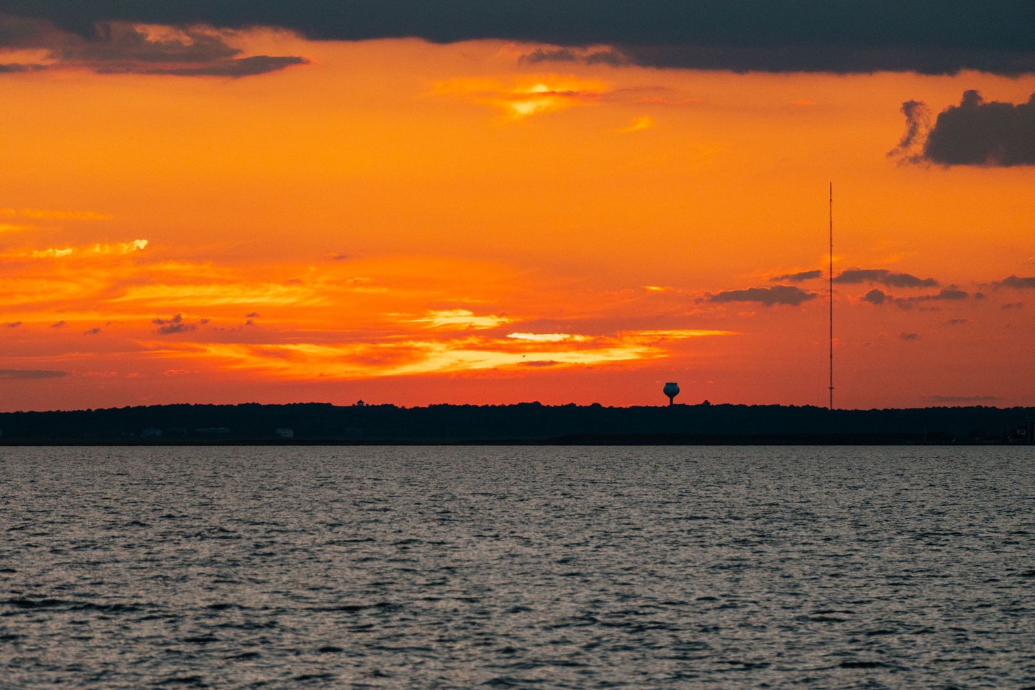 Vivid orange sunset over a lake with silhouetted treeline and a water tower on the horizon.