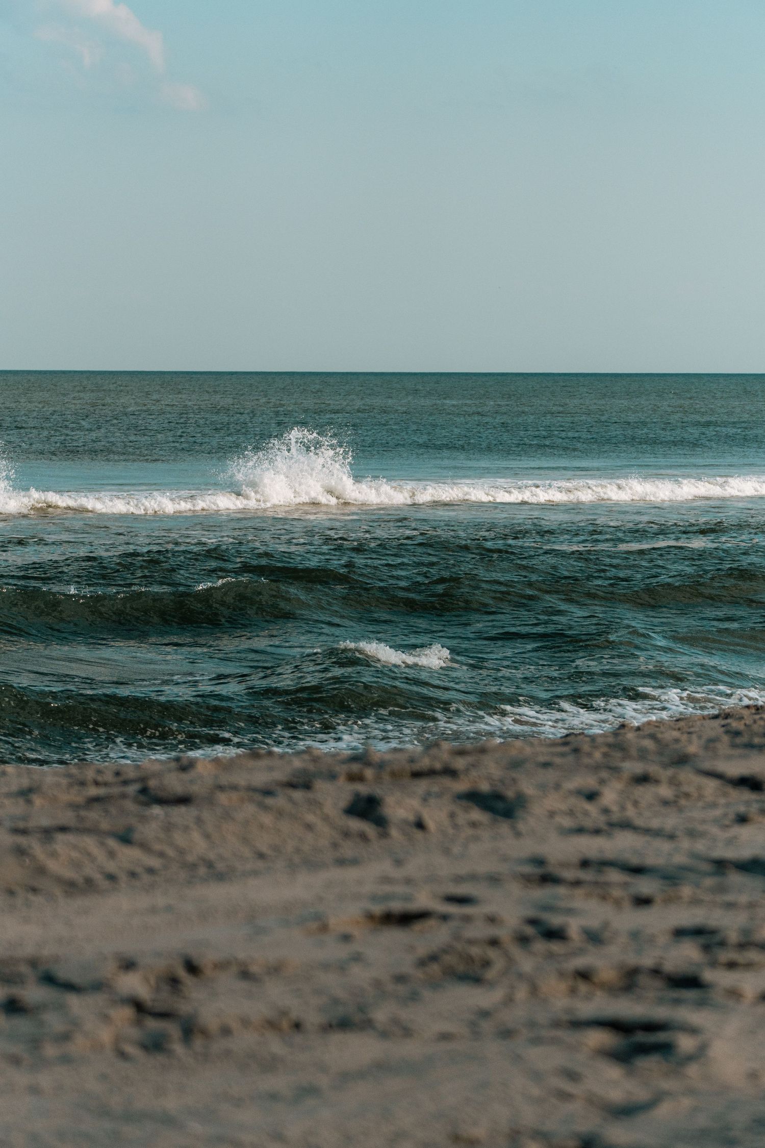 A large wave crashes and splashes on a sandy beach with deep blue ocean water under a clear sky.