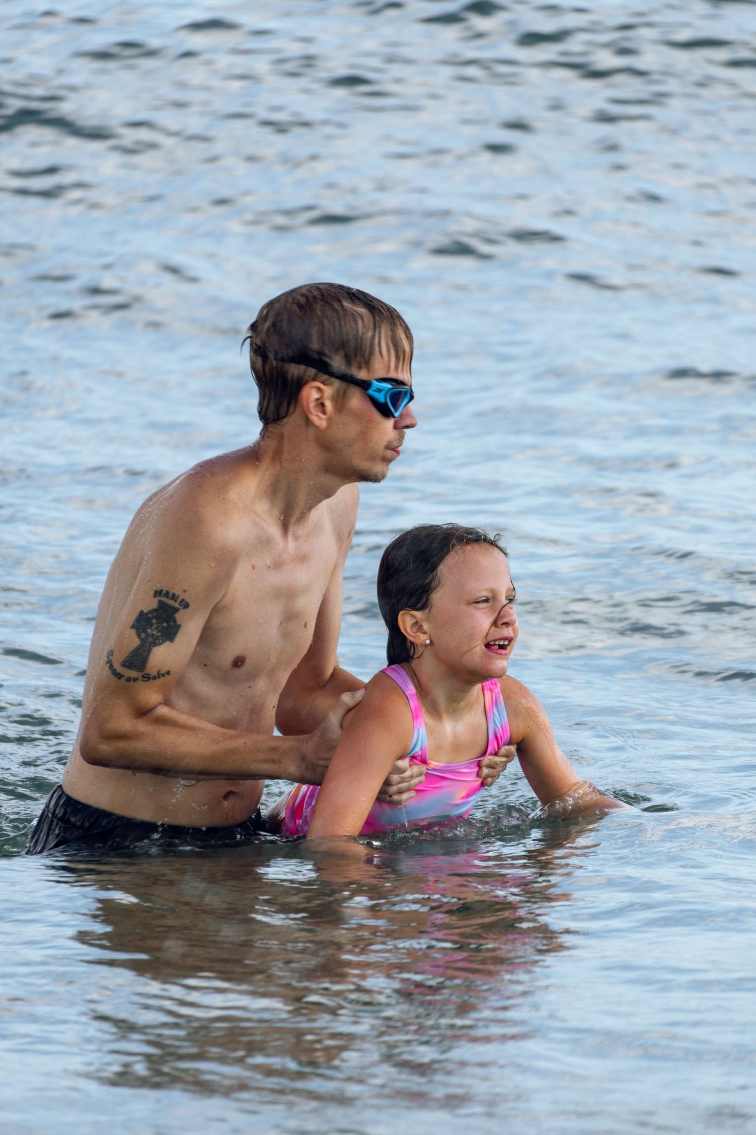 A tattooed man in swim goggles holds a crying young girl in a pink swimsuit in the ocean.
