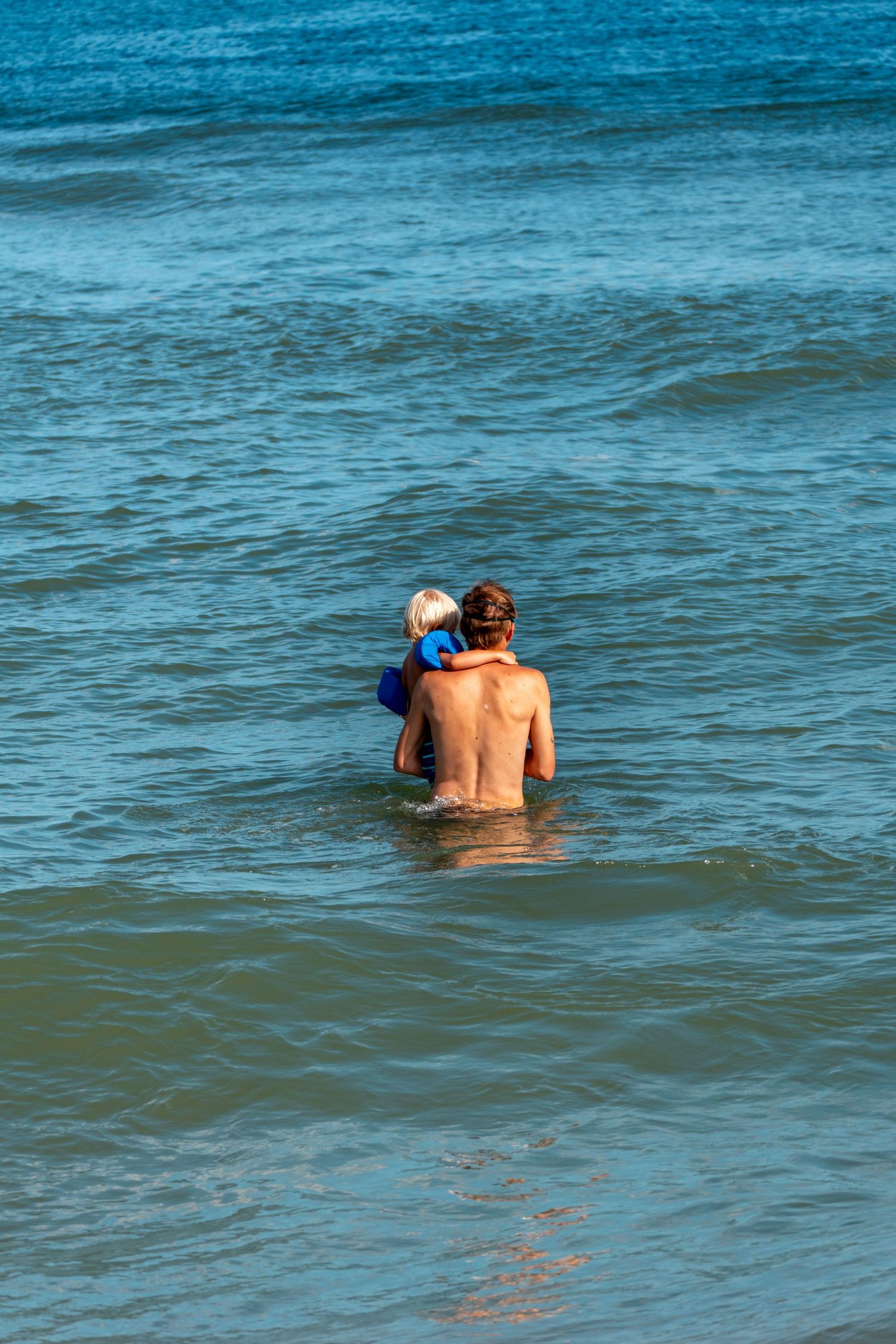 Father carrying young child in blue floaties while wading into blue ocean waves.