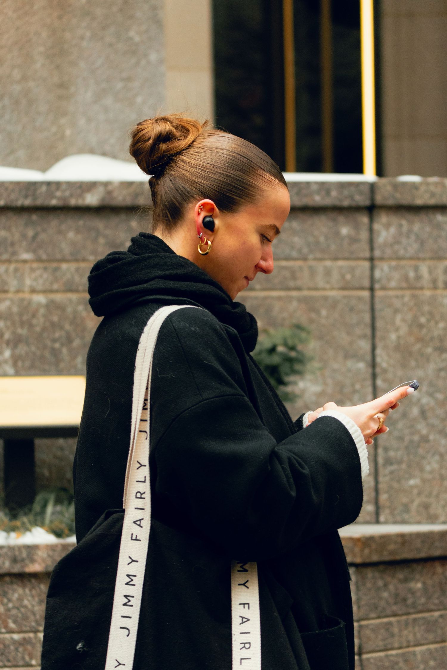 Young woman with hair bun checking phone wearing black Jimmy Fairly coat outdoors.