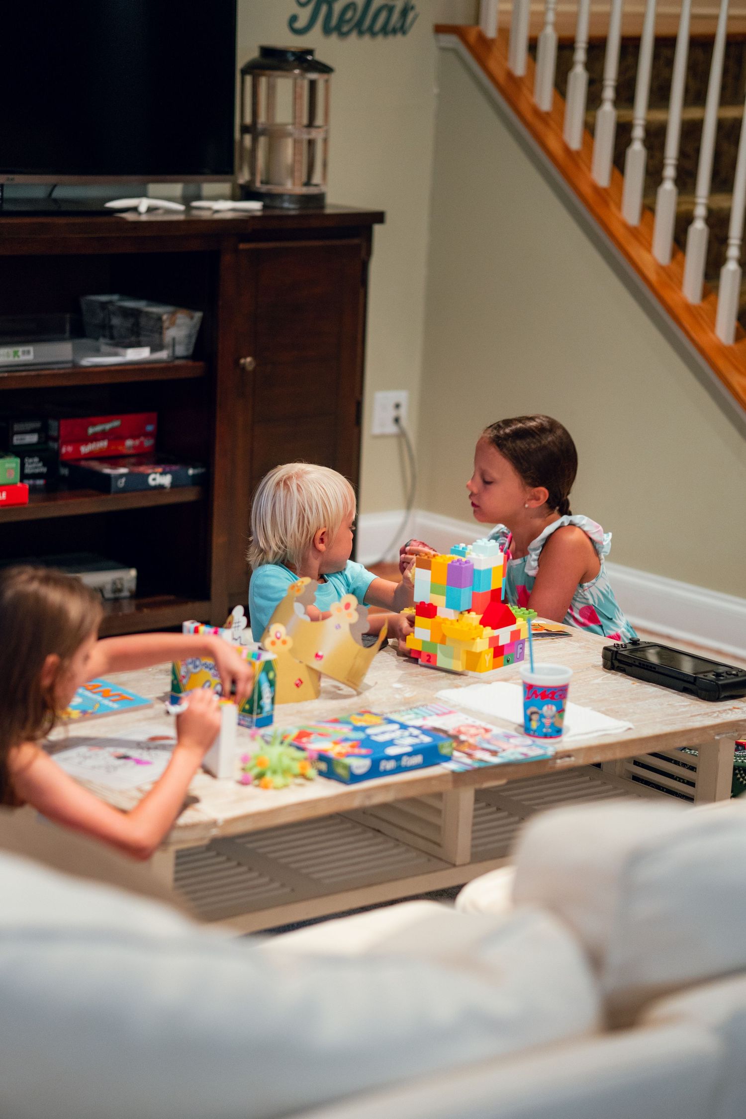 Three children gathered around a coffee table playing with colorful Duplo blocks in a home living room.