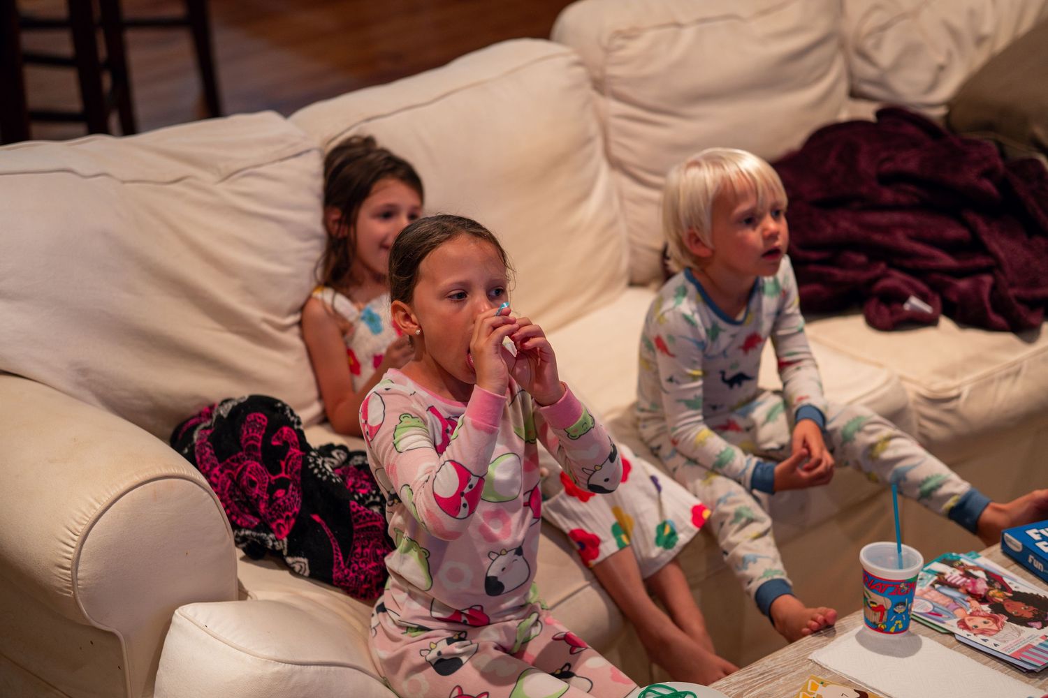 Three children in pajamas sitting on a white couch watching TV together.