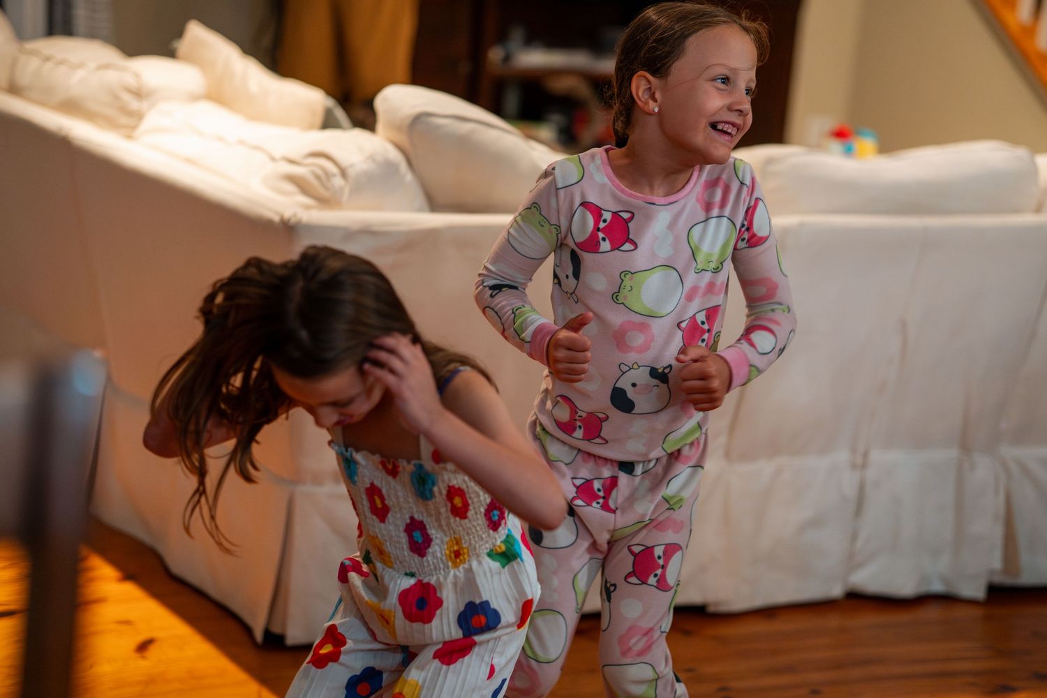Two young girls in colorful pajamas laugh and play together on a hardwood floor at home.