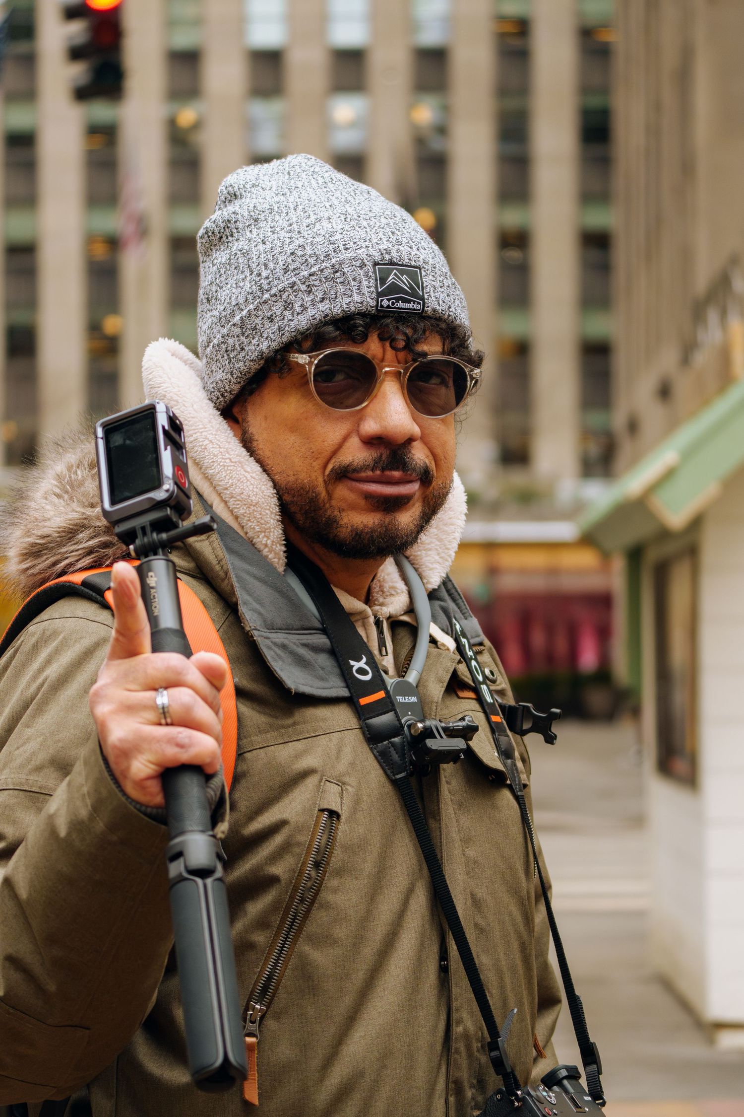 Man in gray beanie and sunglasses holding a GoPro selfie stick on a city street in New York.