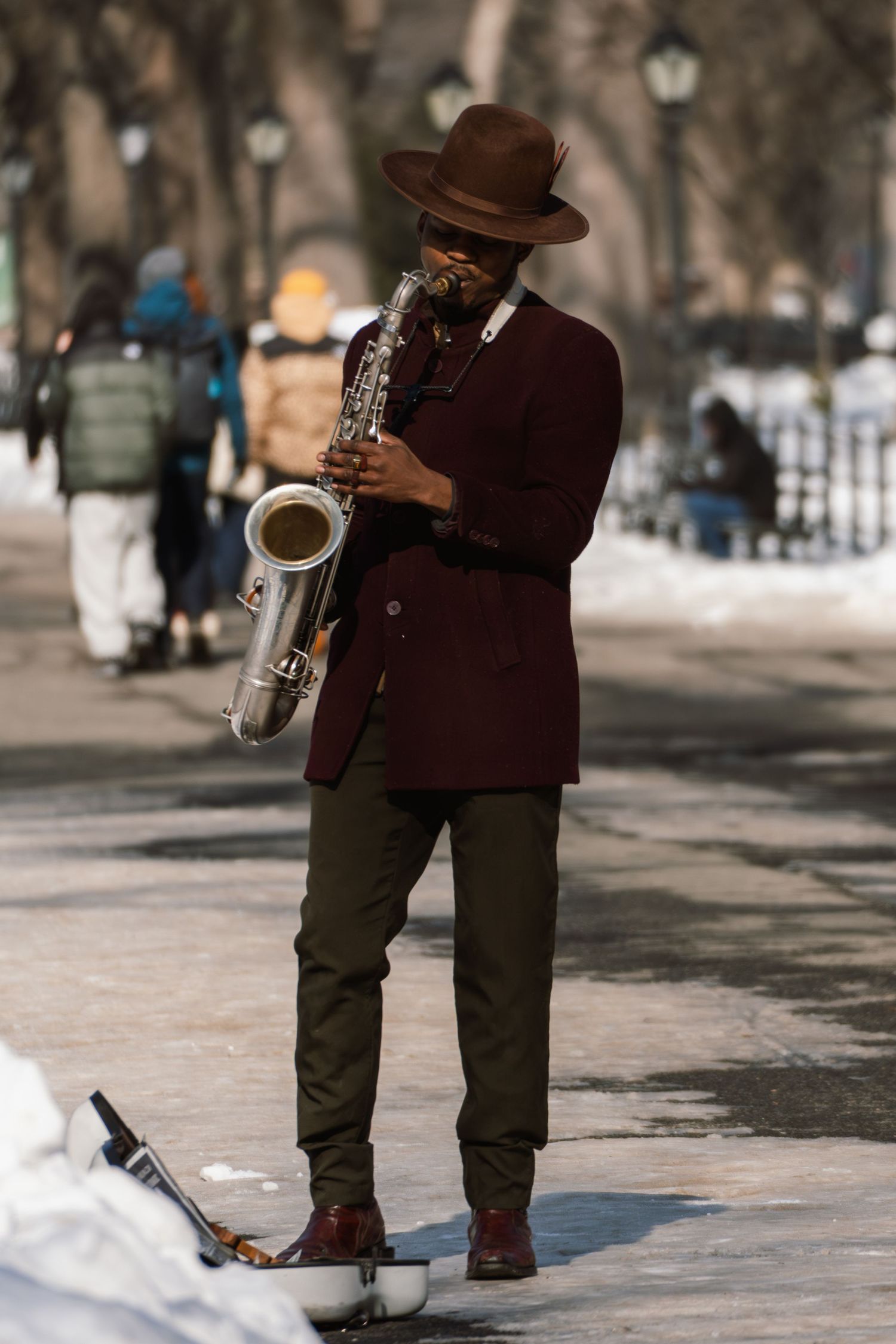 A jazz musician in a burgundy blazer and brown hat plays saxophone on a snowy park path.