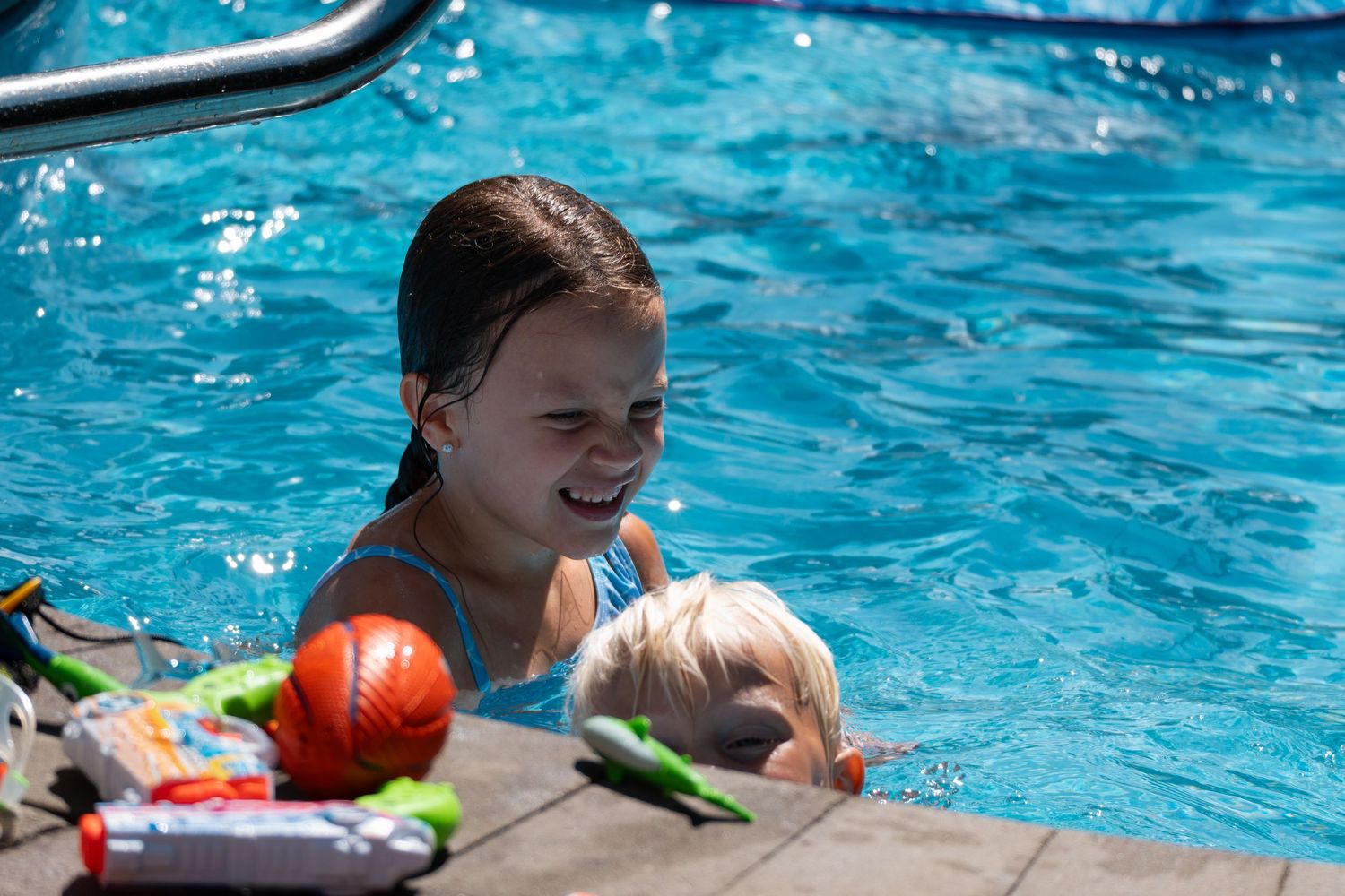 Laughing girl with wet hair in a pool with a young blonde child and colorful toys nearby.
