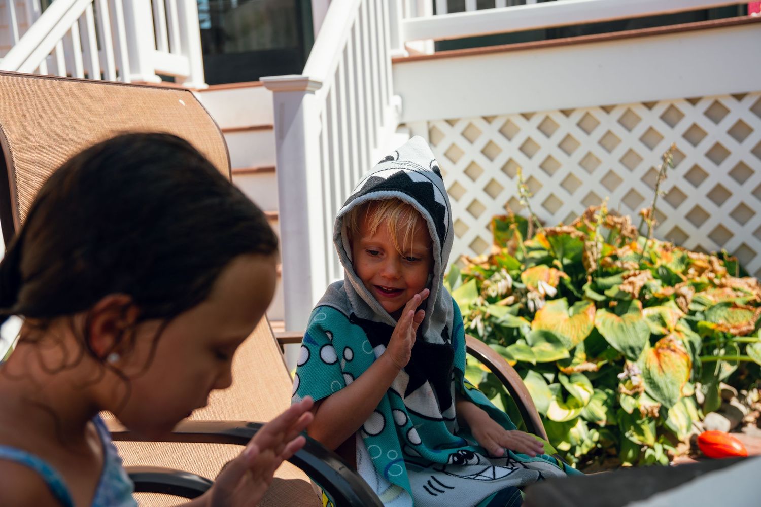Two young children on a sunny deck, one wearing a shark hooded towel poncho, smiling.