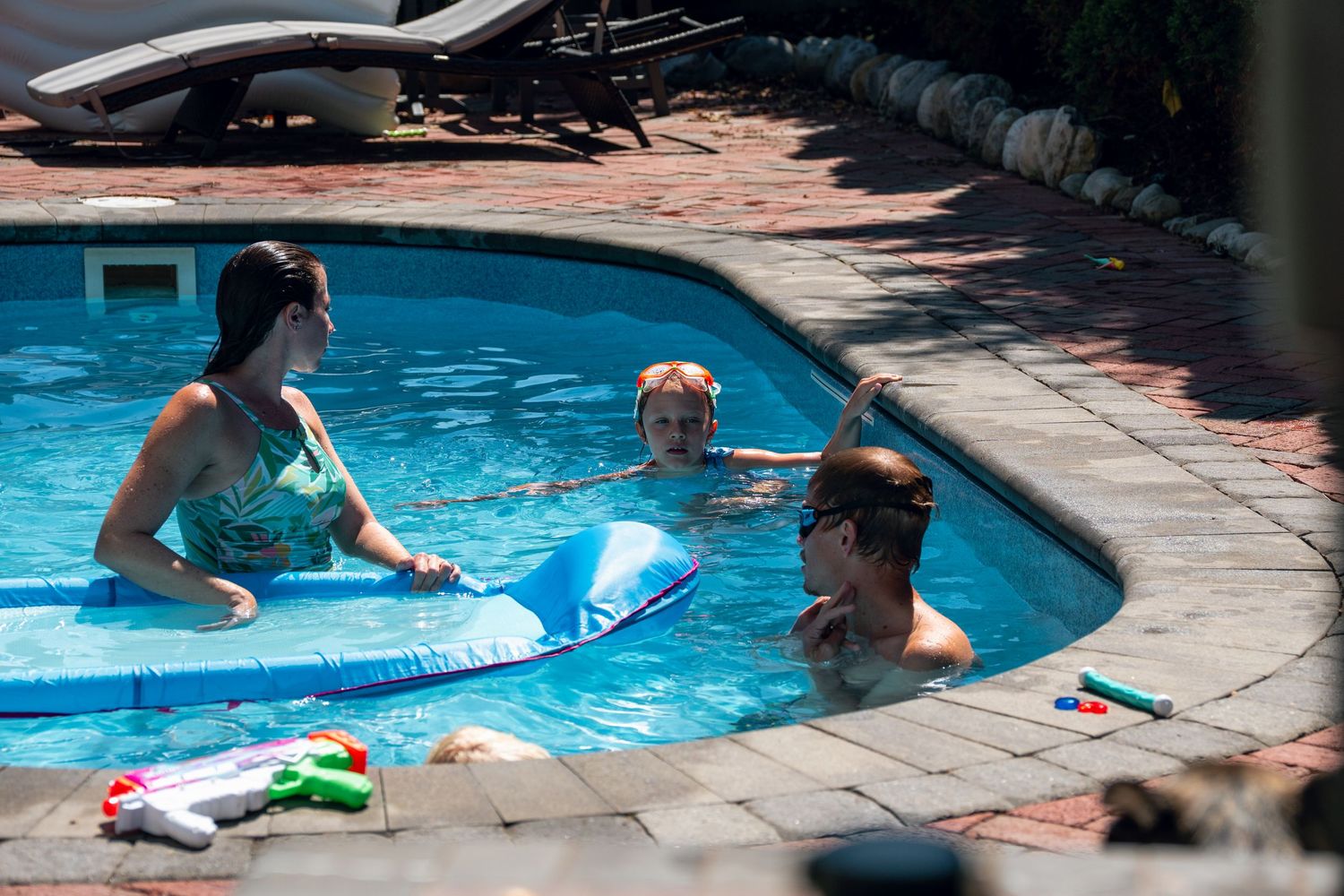 Three people swimming in a backyard pool with water toys on a sunny day.