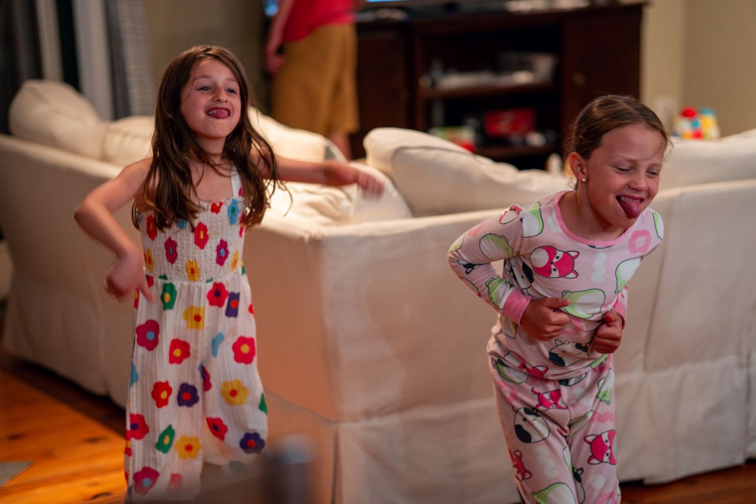 Two smiling girls in colorful pajamas dance and play together on a hardwood floor.