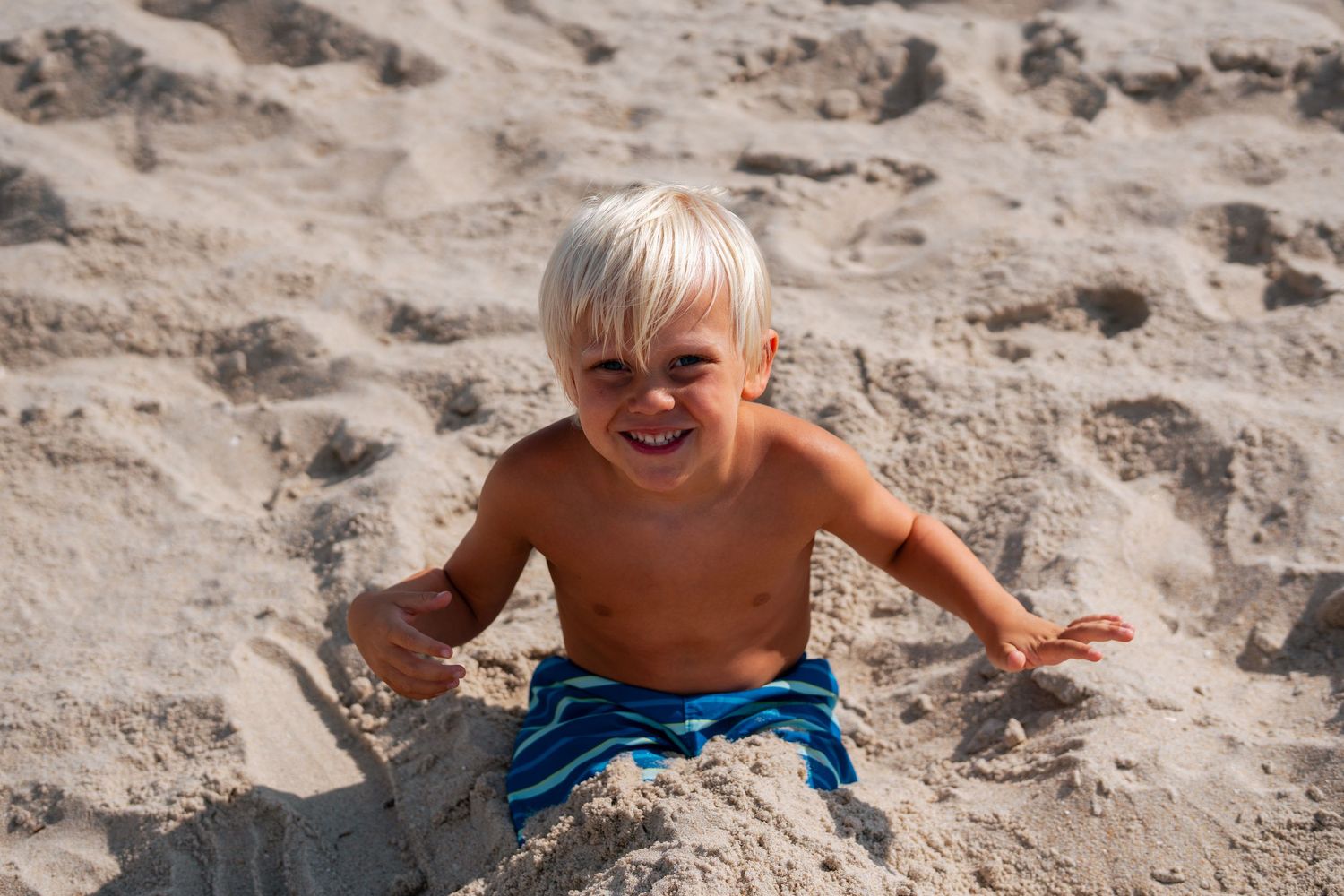 Joyful blonde toddler boy in blue swim trunks laughing while playing in beach sand.