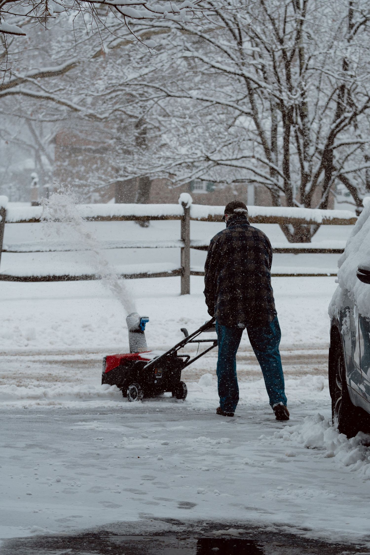 Man using a red snowblower to clear a snow-covered driveway during a heavy winter snowstorm.