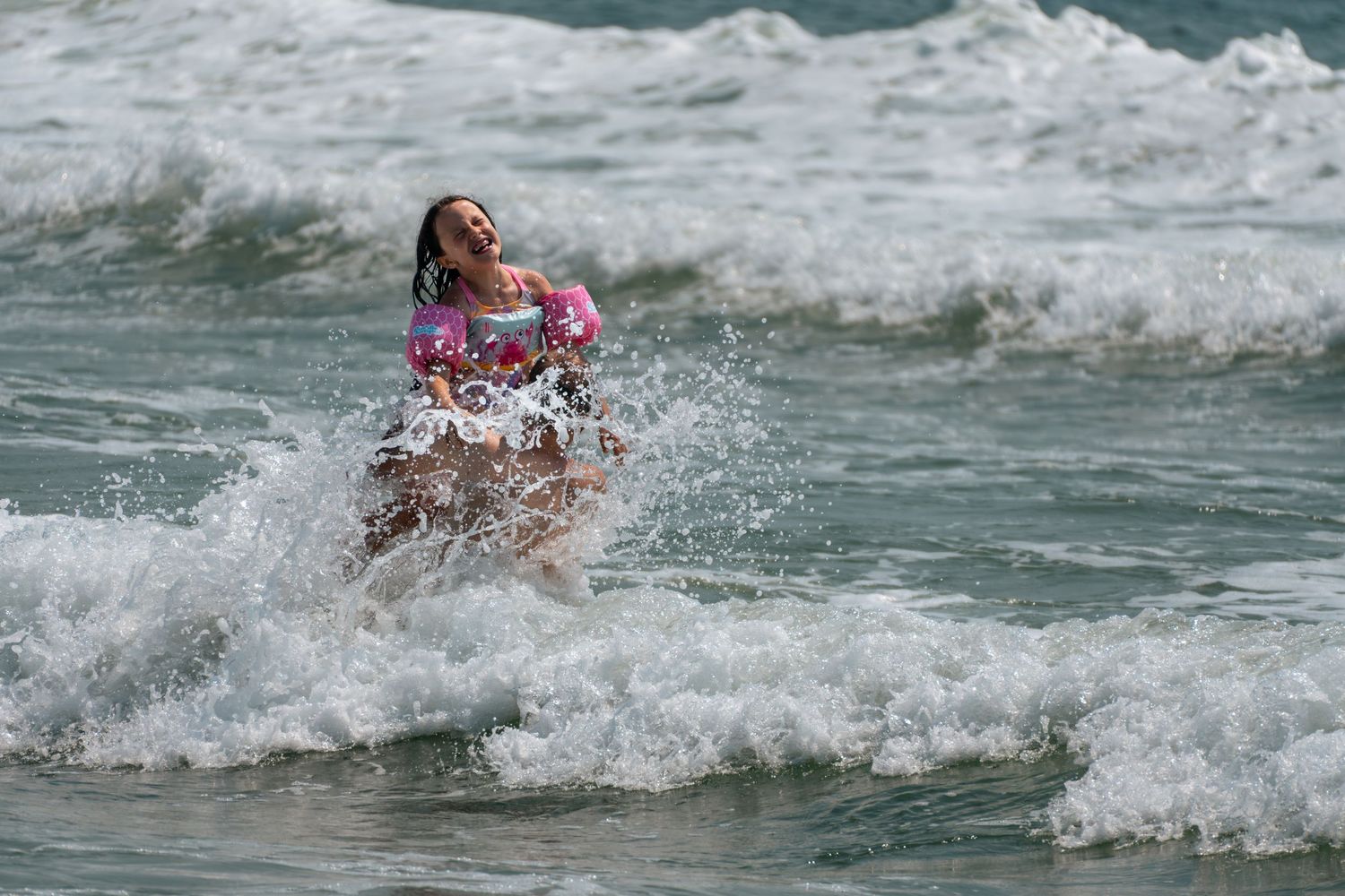 Young girl laughing and playing in ocean waves at the beach, wearing a pink swimsuit and arm floaties.