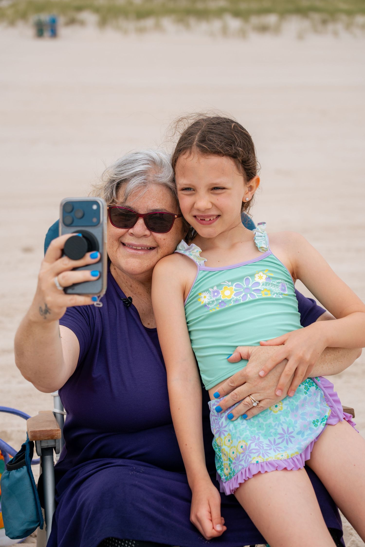 Grandmother and granddaughter taking a selfie at the beach, girl in teal swimsuit, woman in sunglasses smiling.