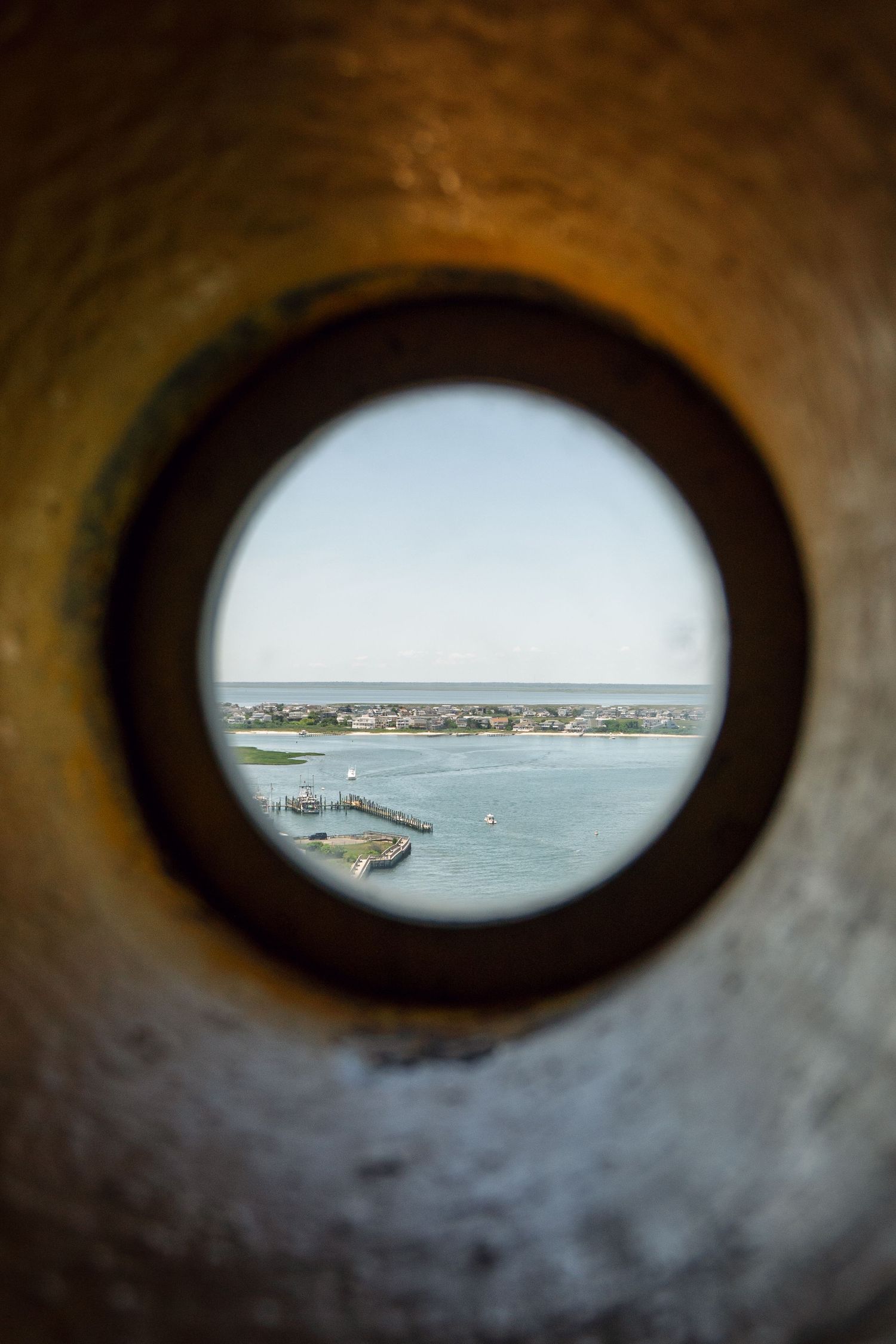 Coastal harbor view through a circular porthole window in a stone wall, showing a pier and distant shoreline.