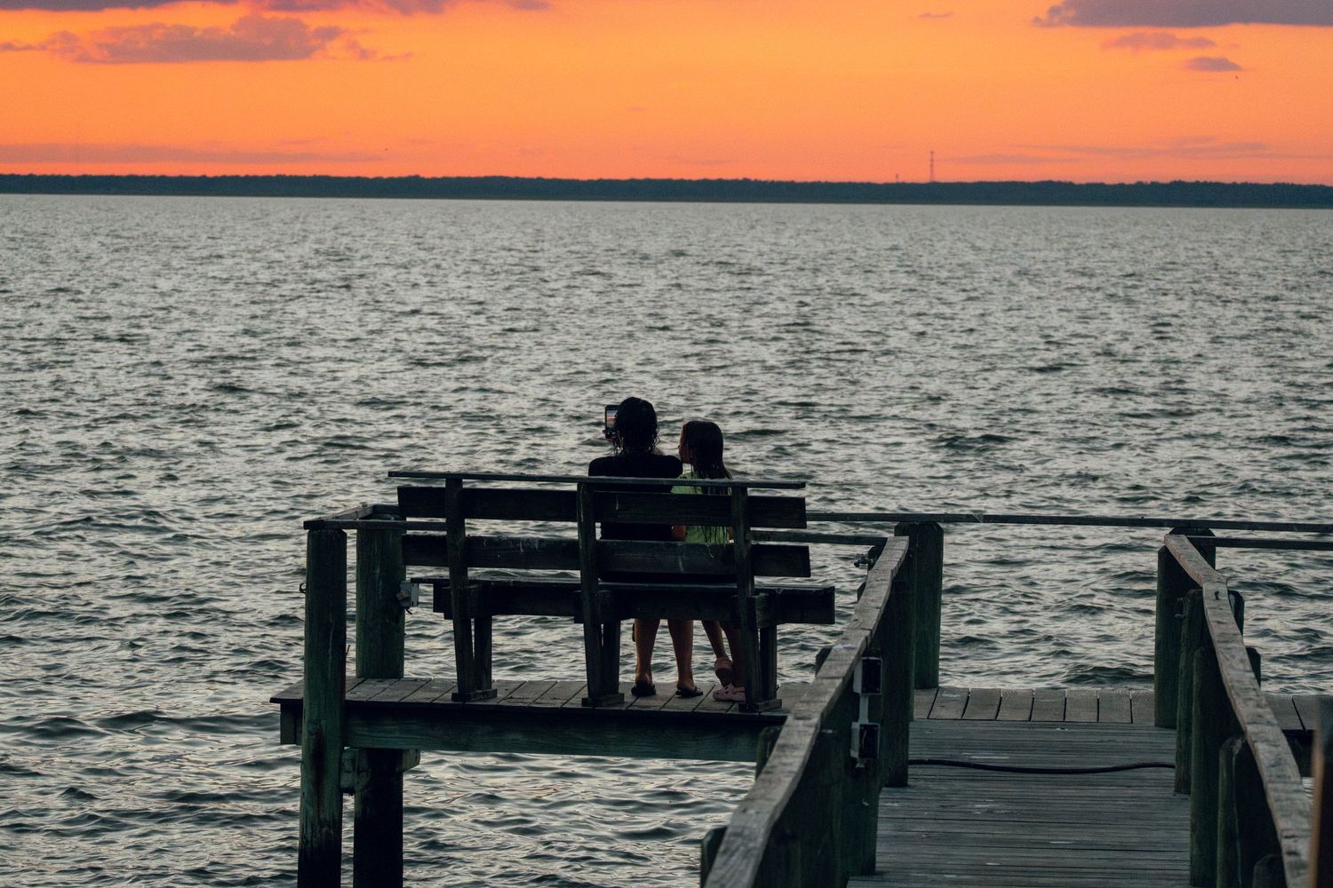 Couple sitting on a wooden dock bench watching a colorful sunset over the bay.