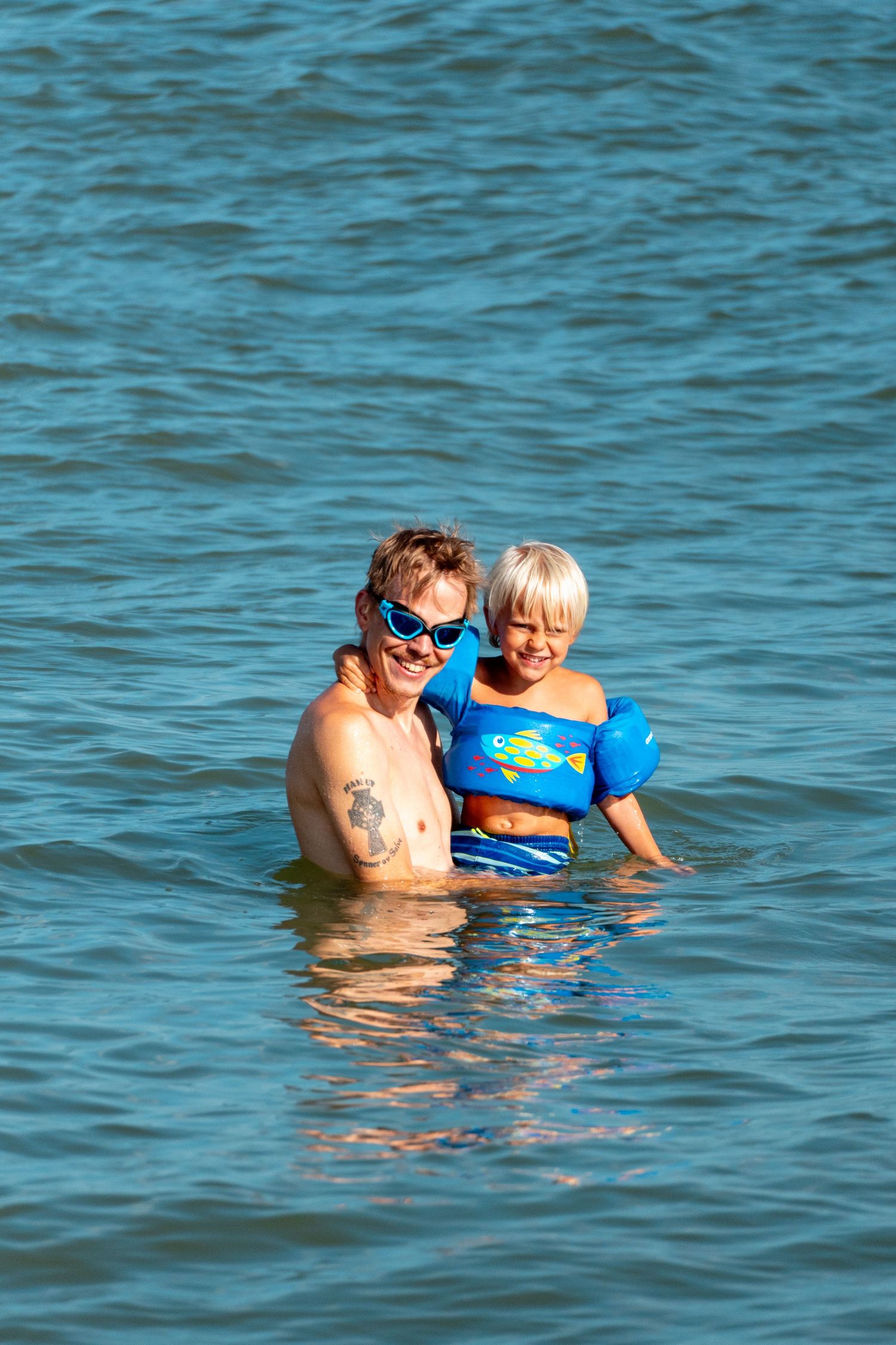 Smiling adult with tattoo and goggles hugging a child in a blue life vest in the ocean.