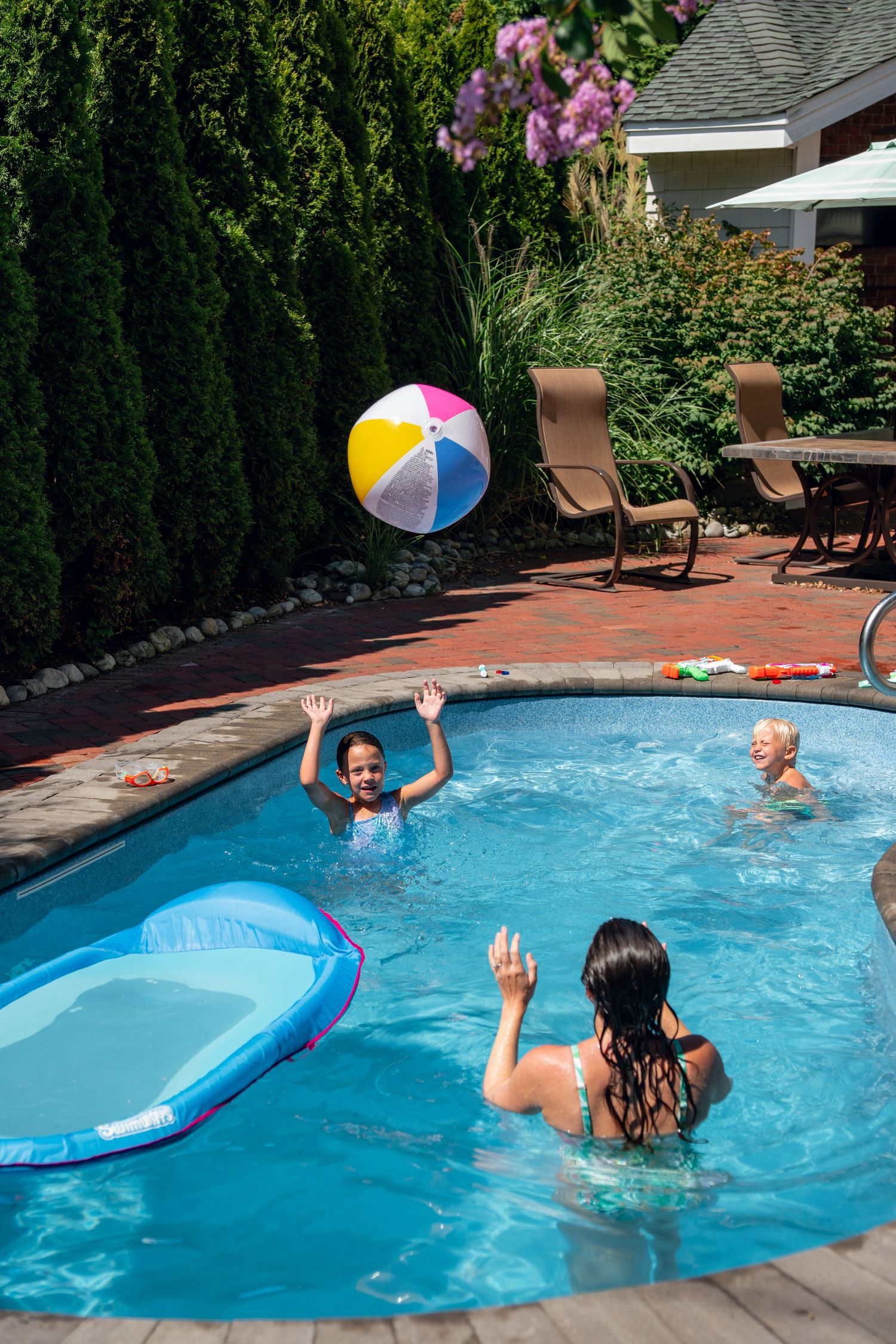 Kids tossing a colorful beach ball in a backyard pool surrounded by patio furniture and lush greenery.