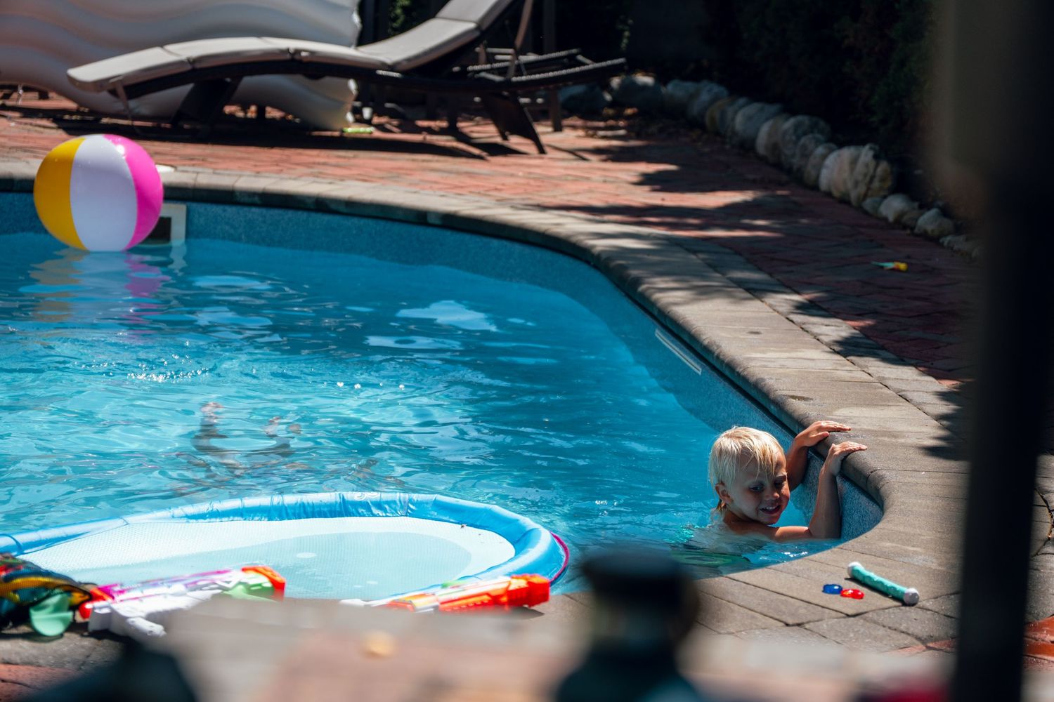 Young child in a backyard pool near a colorful inflatable ring and floating beach ball.