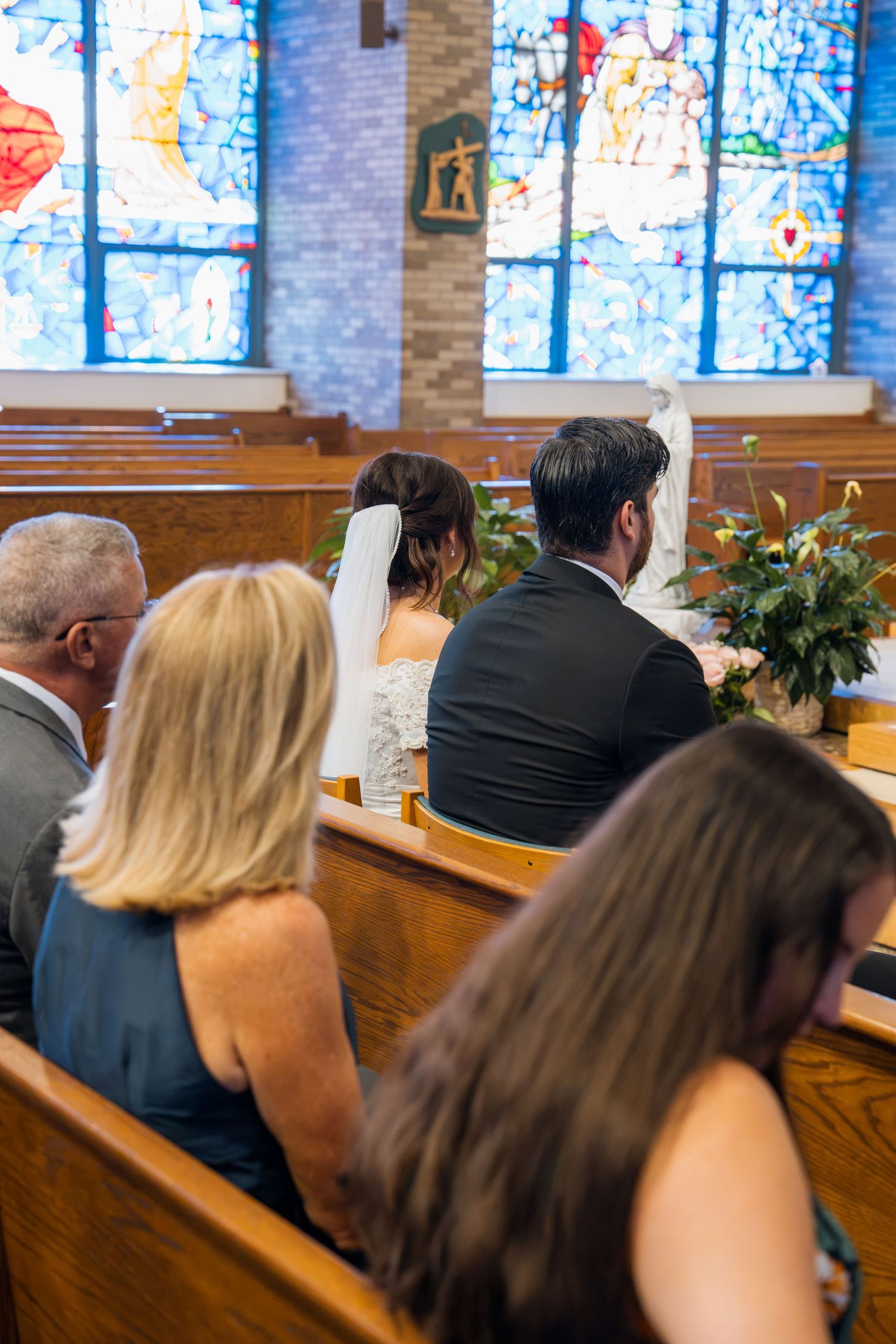 Bride and groom kneeling at church altar during wedding ceremony with stained glass windows and guests seated in pews.
