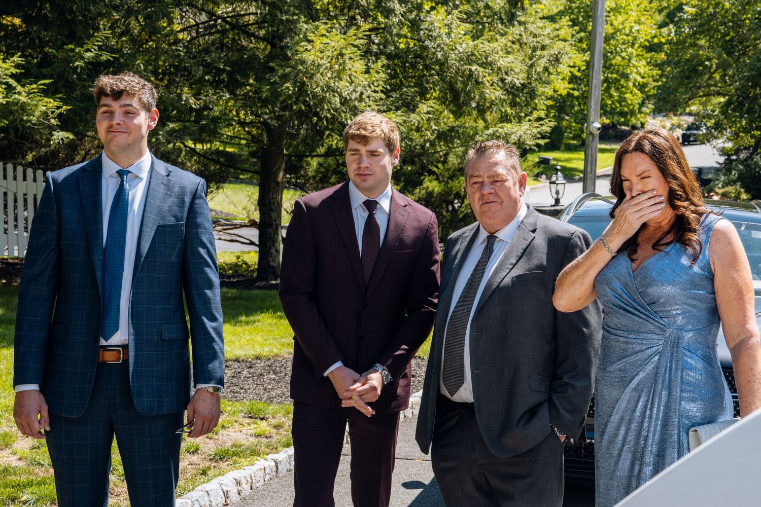 Four formally dressed people posing outdoors at a sunny event, two young men in suits and an older couple smiling.