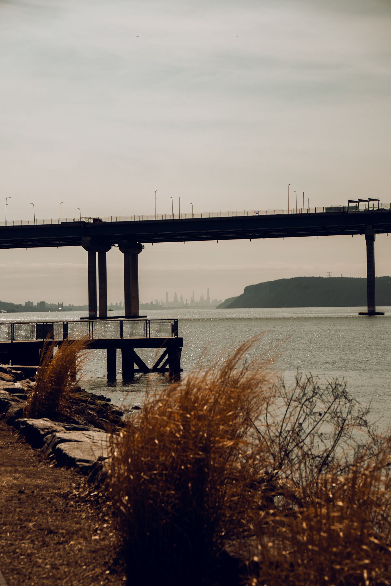 Bridge over calm river with tall grass in foreground under overcast sky.