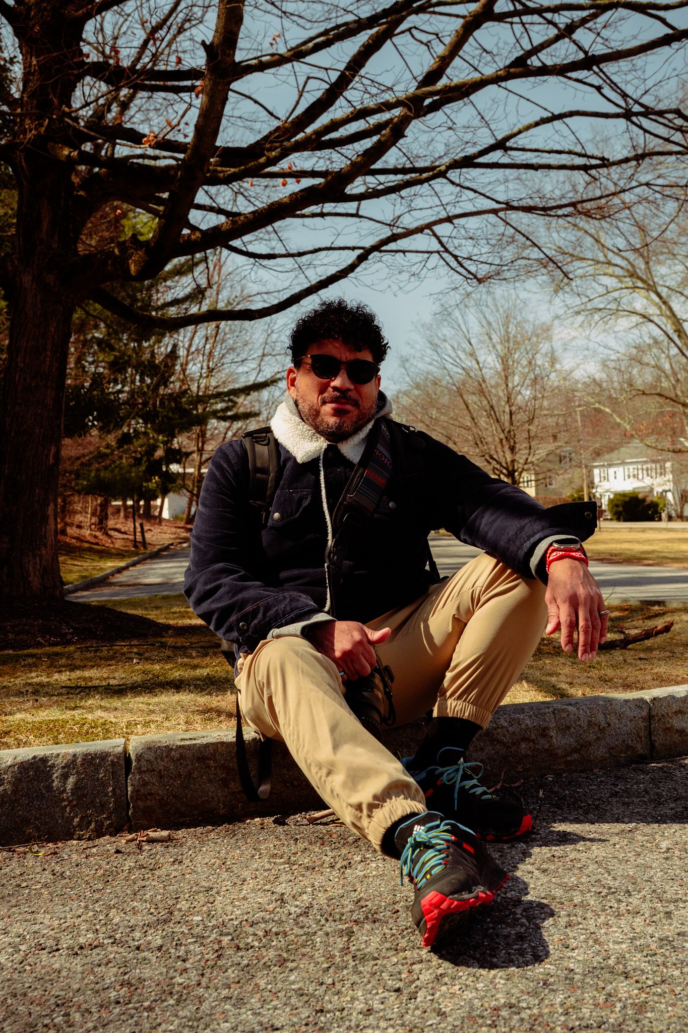 Stylish man in sunglasses and navy jacket sitting on a stone curb in a sunny winter park.