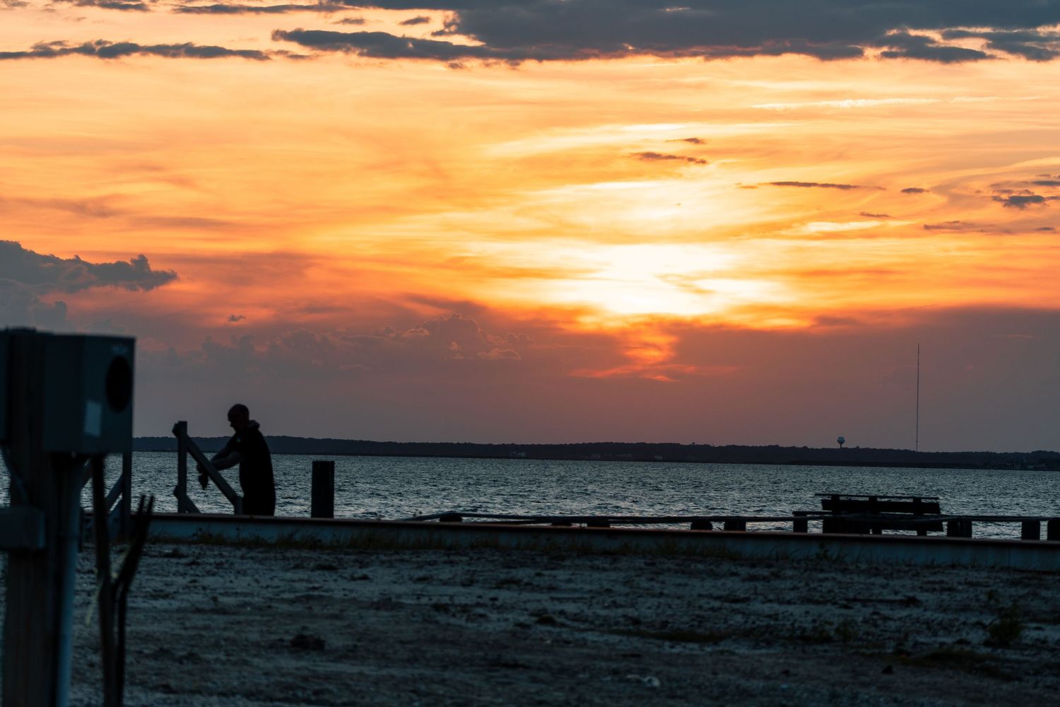Silhouette of a person on a dock during a vibrant orange and golden sunset over calm ocean waters.
