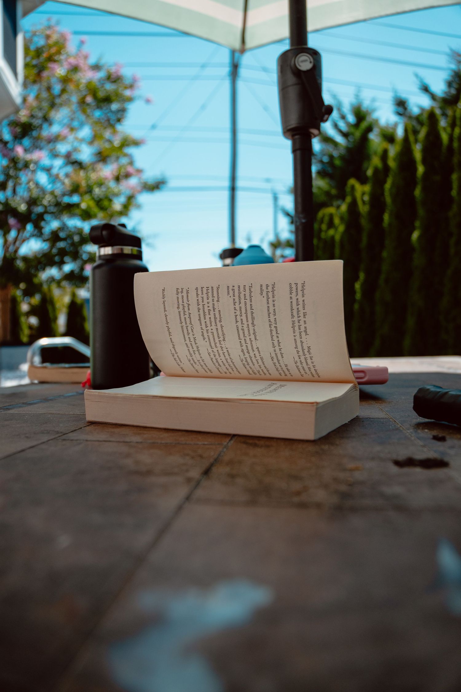 Open book on a tiled patio table with a thermos beside a sunny backyard pool.
