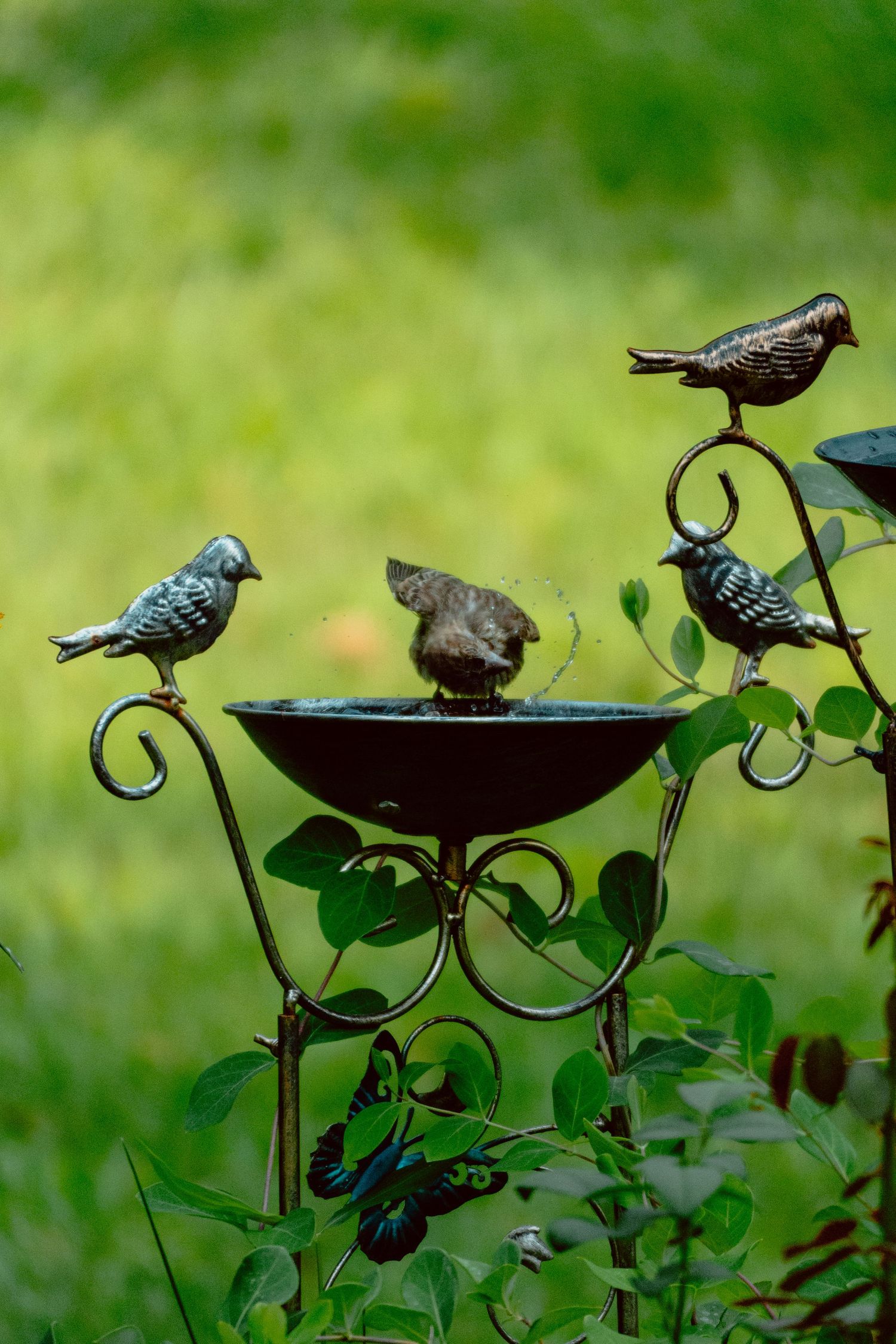 A small bird splashes in a decorative metal bird bath with ornamental bird sculptures on a green garden background.