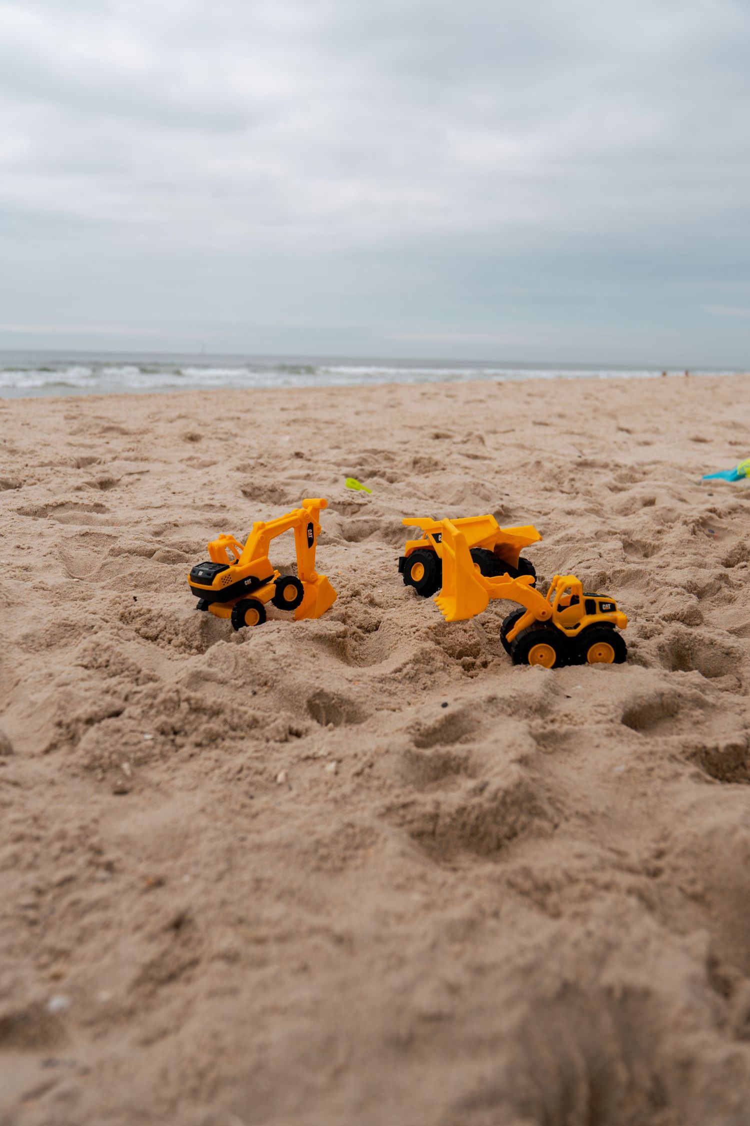 Yellow toy construction vehicles playing in sandy beach with ocean waves and cloudy sky in background.