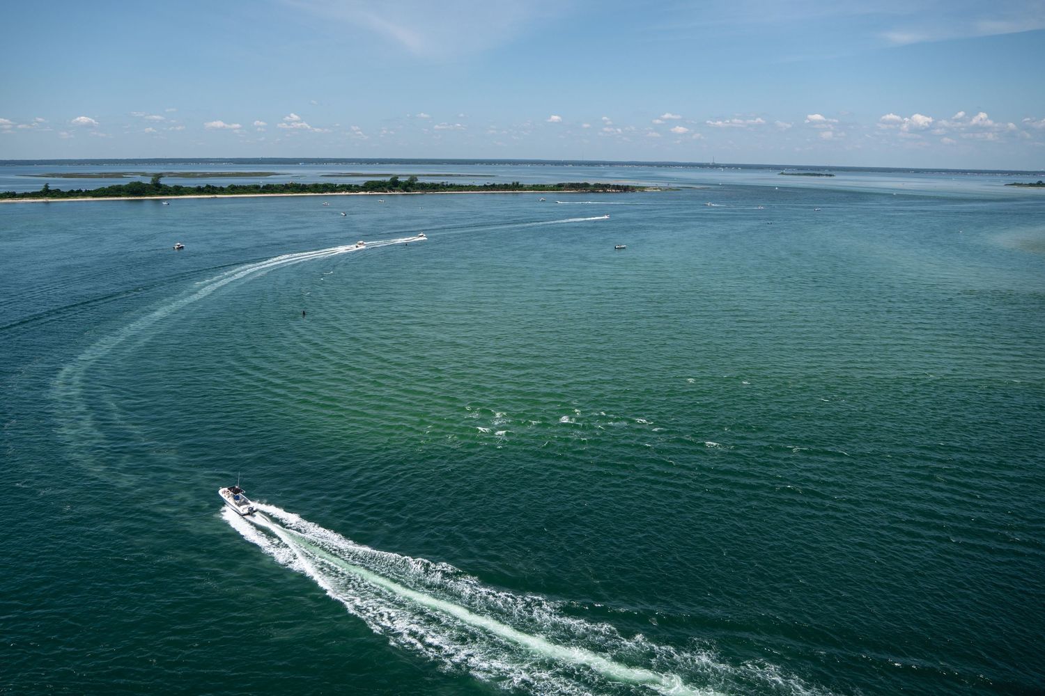 Aerial view of a speedboat creating rippling wake patterns on calm turquoise waters near a small island under blue skies.
