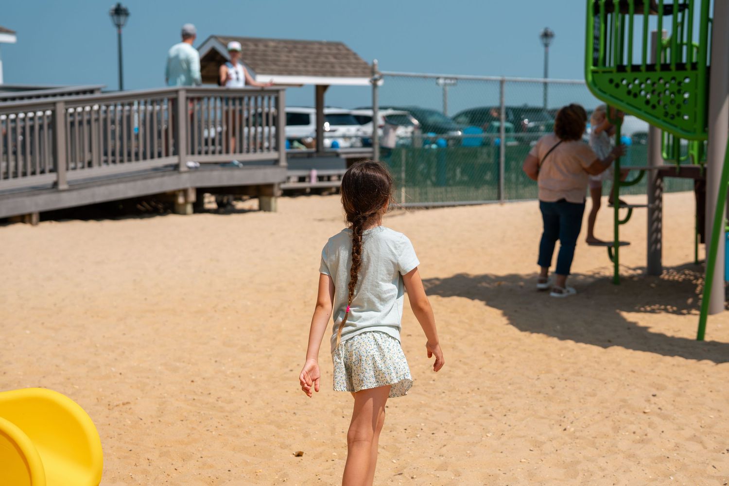 A young girl with a braid walks toward a playground with green equipment on a sandy surface near a boardwalk.