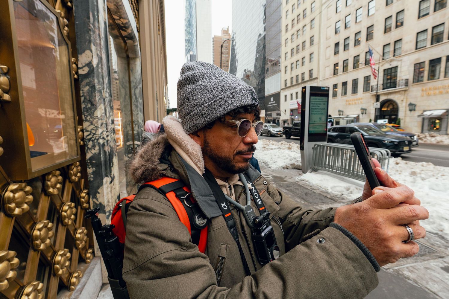 Man with sunglasses and gray beanie taking selfie on snowy New York City street.