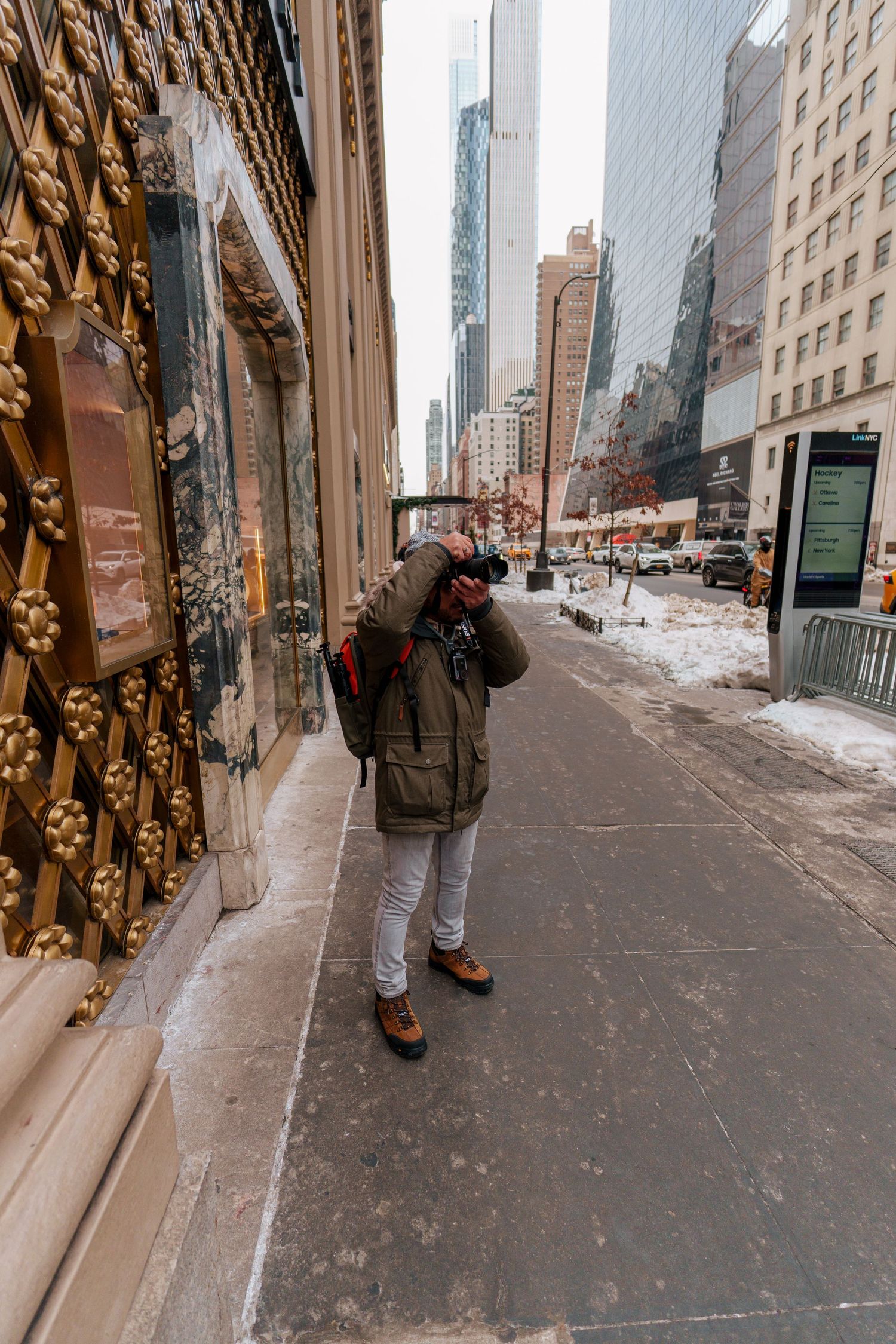 A photographer in a green jacket and beanie captures photos near golden ornate doors on a NYC street.
