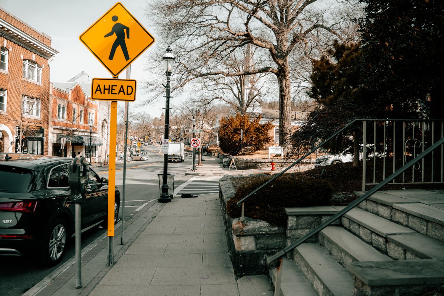 A yellow pedestrian crossing ahead warning sign on a city sidewalk with stairs and bare trees in the background.