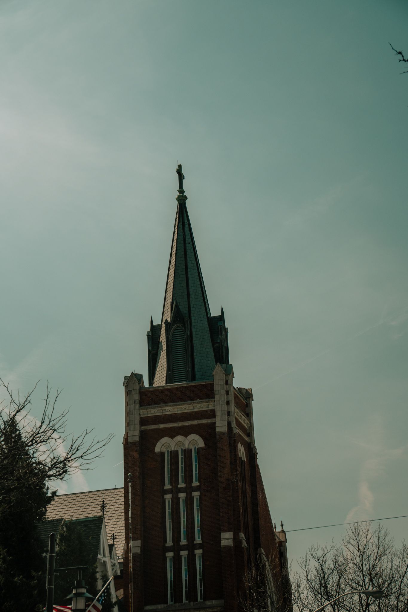 Red brick church steeple with green spire and cross against a cloudy sky in a small town.
