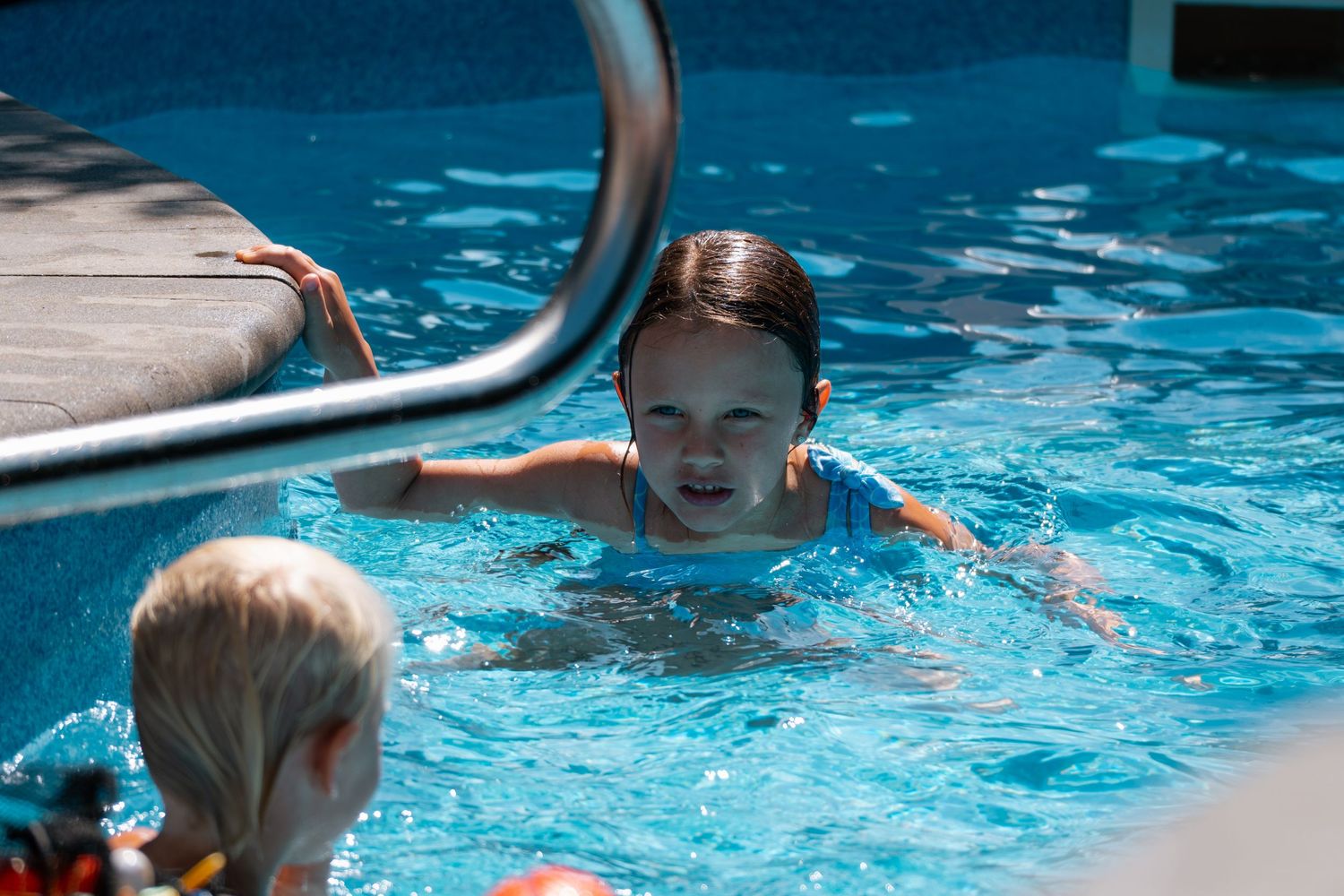 Girl in blue swimsuit swimming in a bright blue outdoor pool near metal ladder.