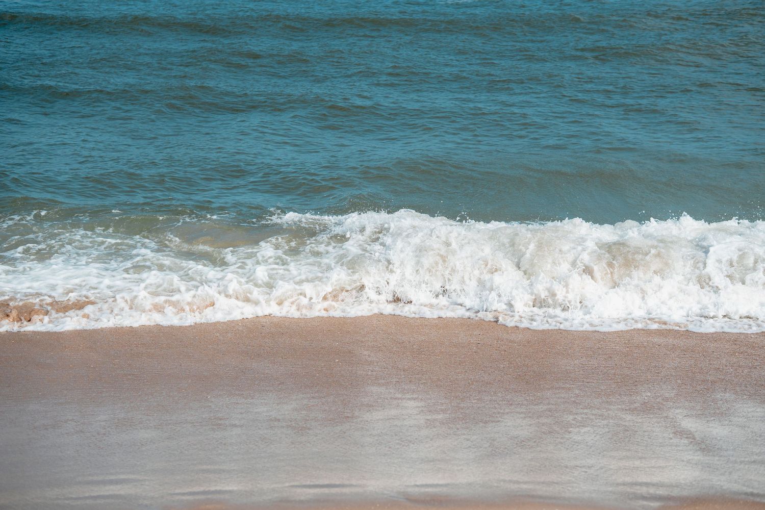 Gentle ocean waves with white sea foam washing onto a smooth sandy beach shore.