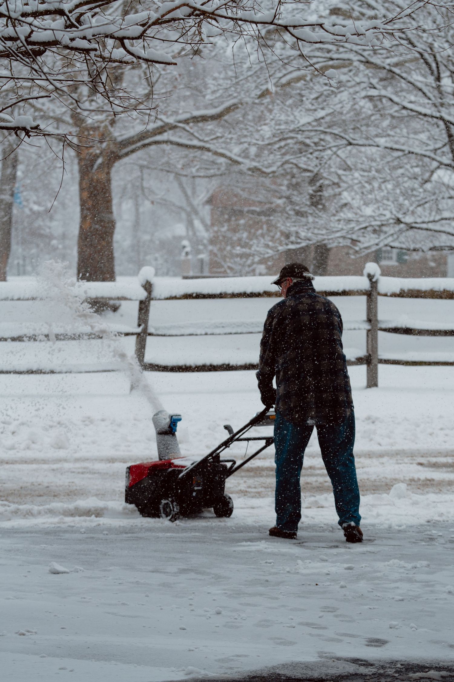Man clearing snow from driveway with a red snowblower as snow falls heavily in winter.