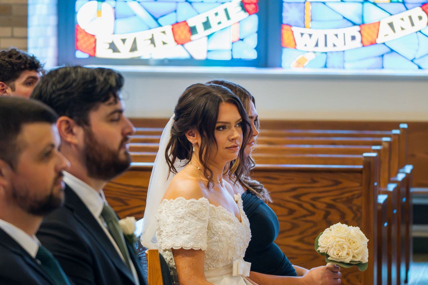 Bride in white lace off-shoulder gown holding bouquet seated in church pew during wedding ceremony.